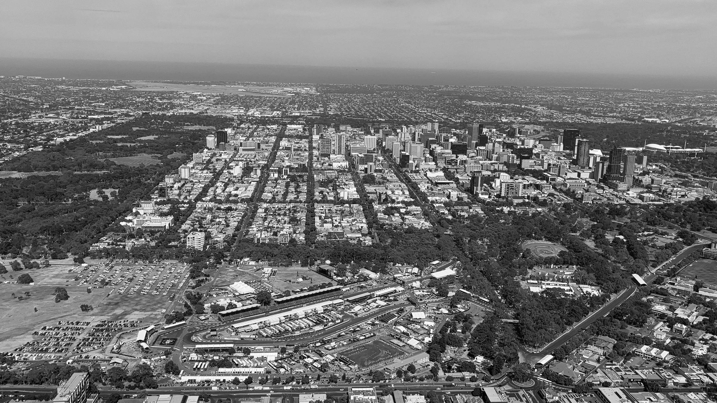 An aerial view over wide parkland dotted with trees, a temporary racing circuit with a pit building and grandstands in the foreground, and the grid of a city centre stretching to the sea beyond.