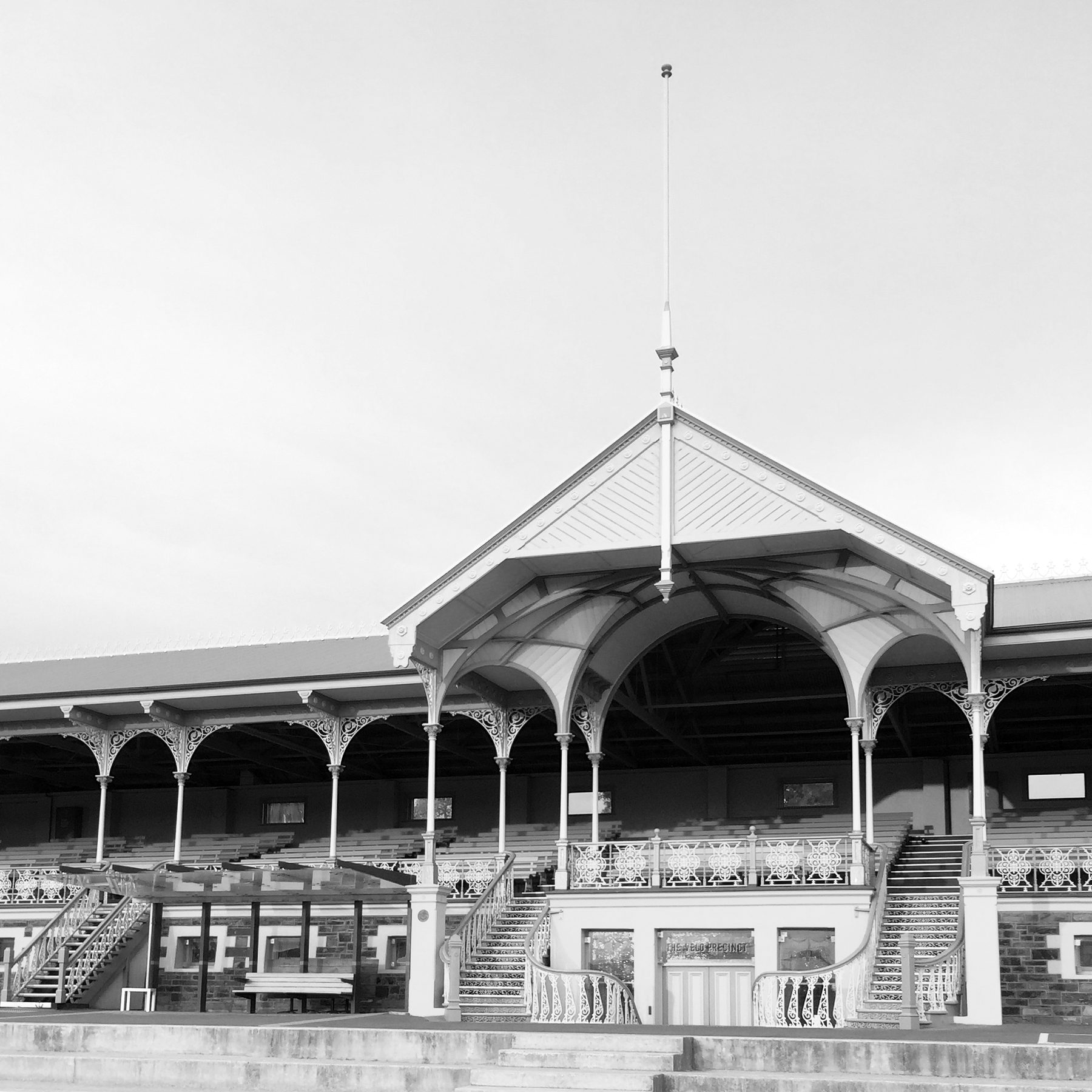 A Victorian grandstand with an ornate iron-lace pavilion roof and a tall flagpole, its tiers of seating empty, seen across a paved forecourt.