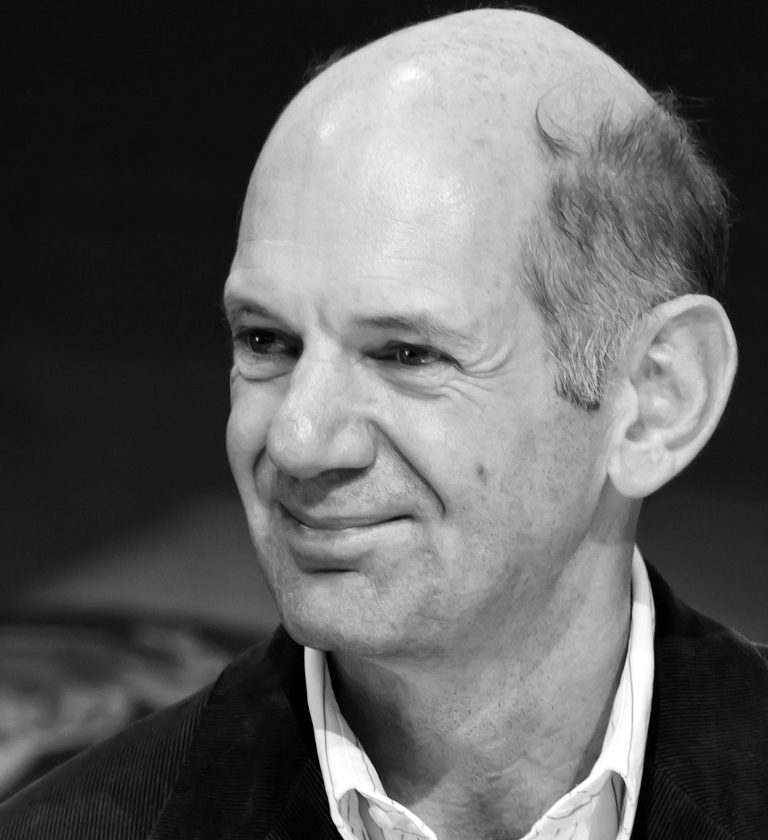 Head and shoulders of a balding man with greying hair at the sides, smiling slightly and looking off to the left, in a corduroy jacket and open-necked striped shirt.