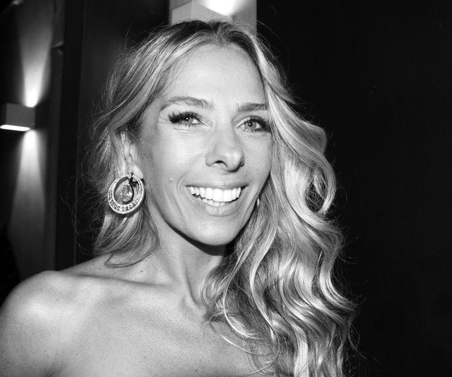A smiling woman with long, wavy fair hair and a large jewelled hoop earring, lit by a flash against a dark wall.