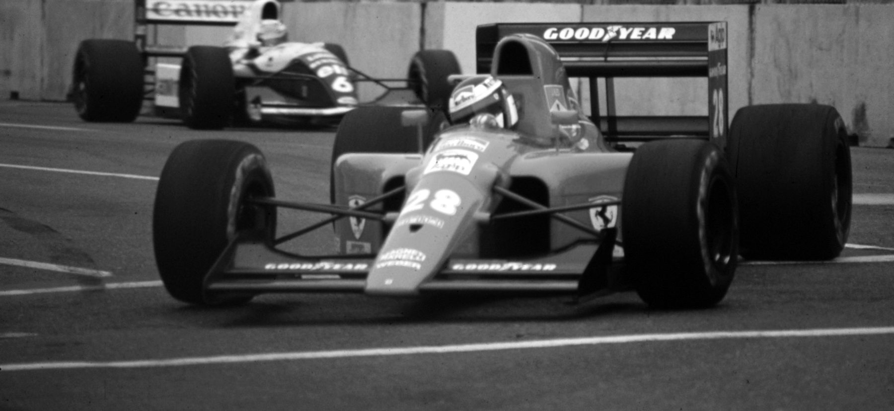A Ferrari, number 28, turns towards the camera on a concrete-walled street circuit, the driver’s helmet just visible above the cockpit; a two-tone Williams, number 6, follows behind.