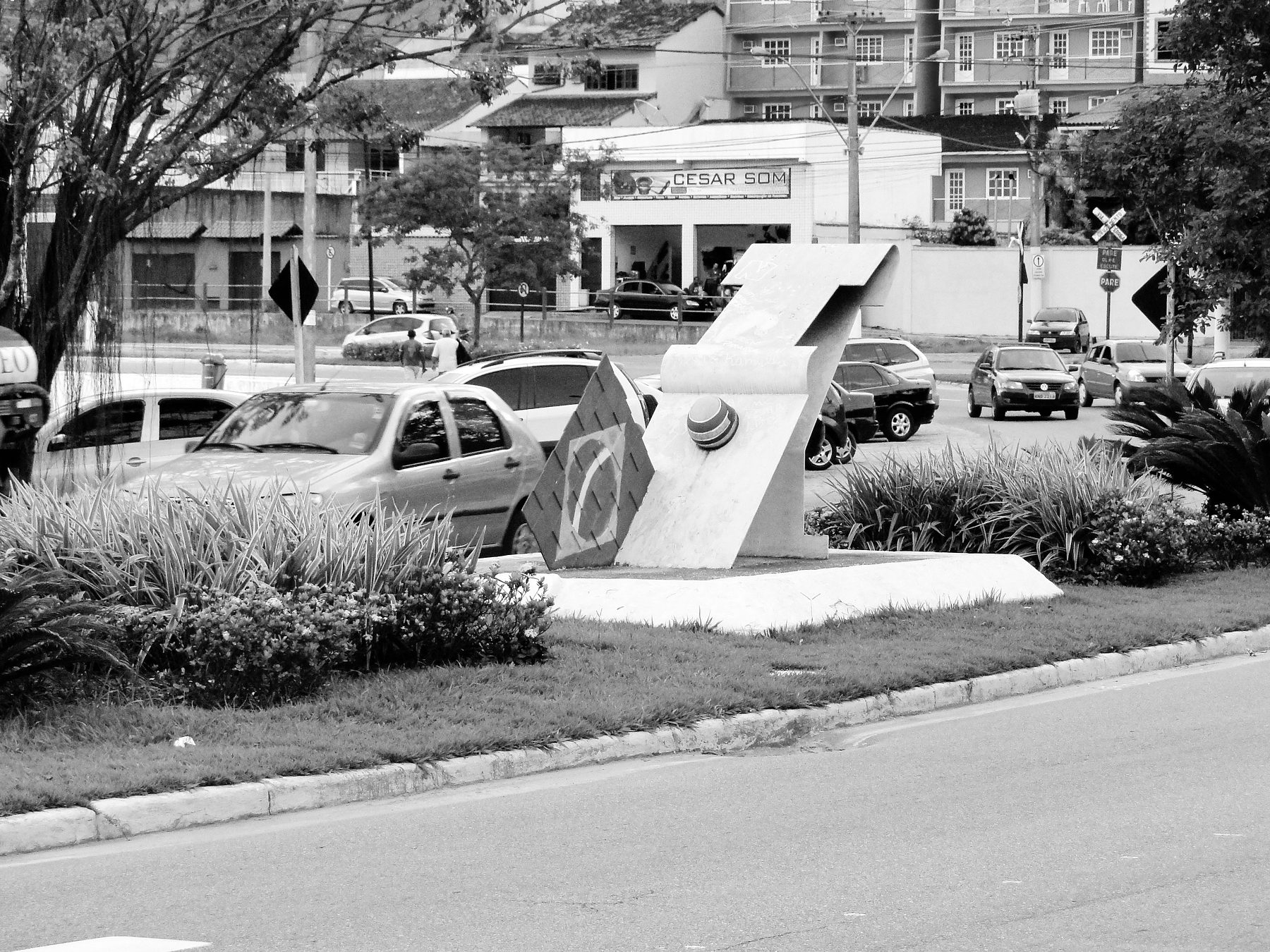 A white concrete monument on a traffic island, a tall form like a raised ribbon of track with the flag of Brazil set into it and graffiti on its surface.