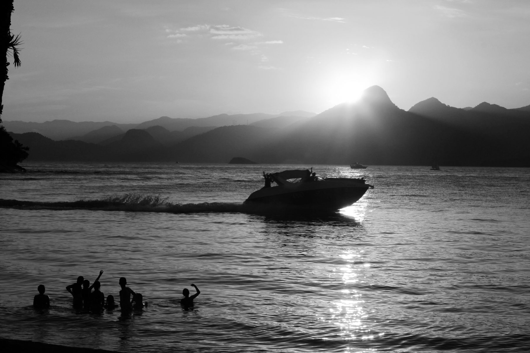 A motor launch cuts across calm water at sunset, the sun setting behind a peaked mountain; swimmers stand silhouetted in the shallows in the foreground.