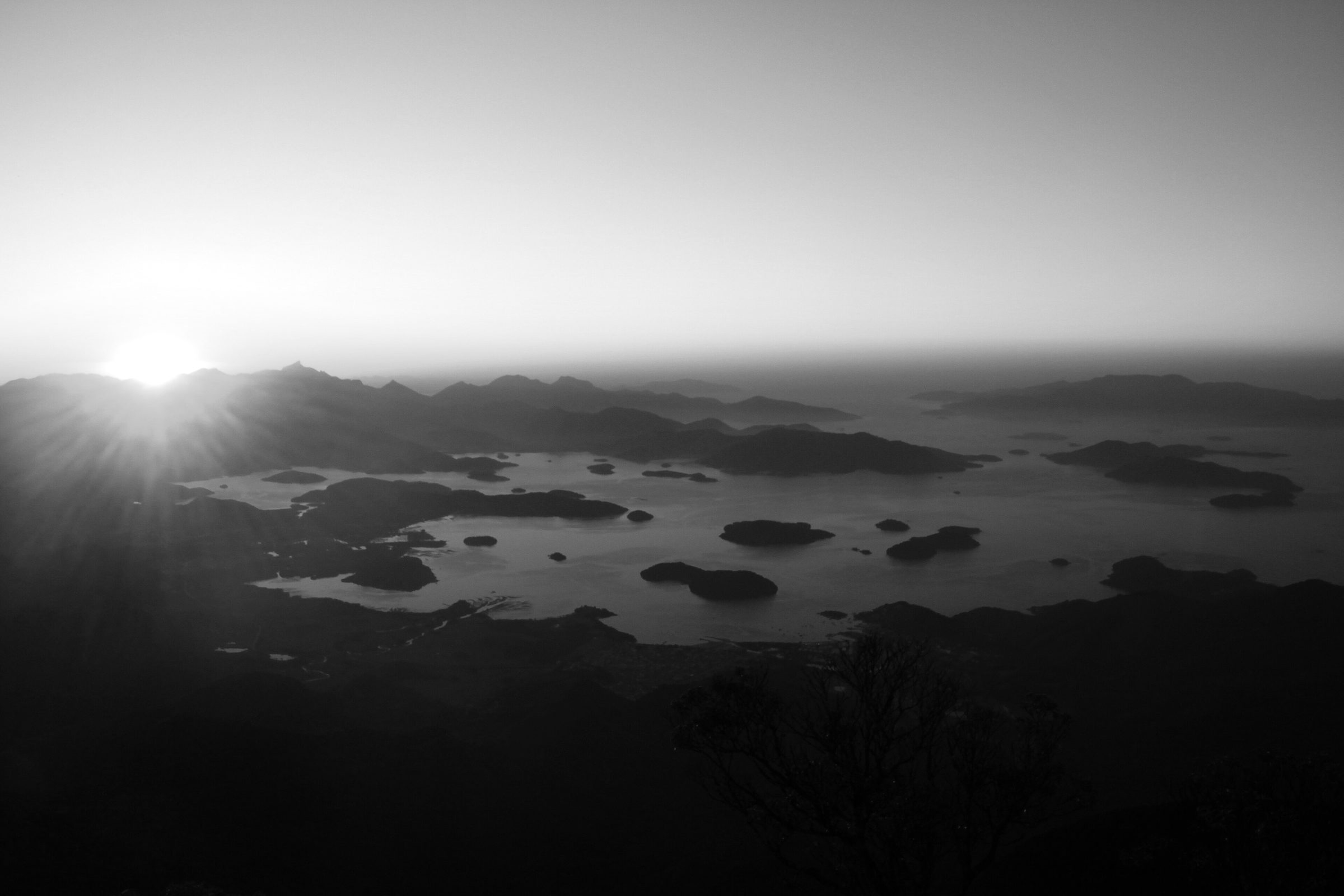 Sunrise from a mountain top over a wide bay scattered with wooded islands, the sun low on the horizon and the far shore in mist.