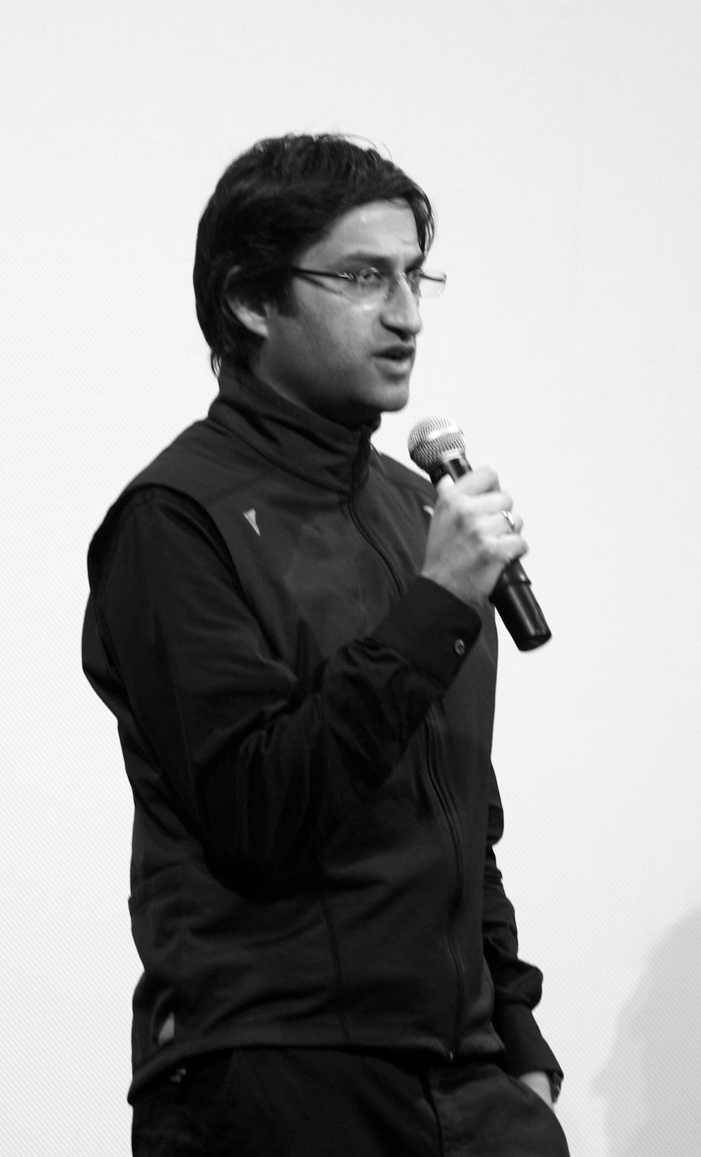 A man in rimless glasses and a dark zip-up jacket speaks into a hand microphone in front of a pale screen.