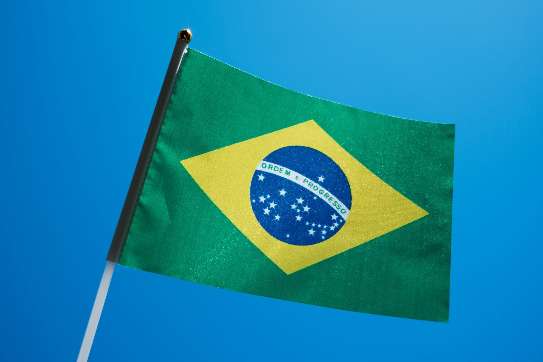 A small Brazilian flag on a thin staff, its green field, yellow diamond and starry blue globe bright against a clear blue sky.