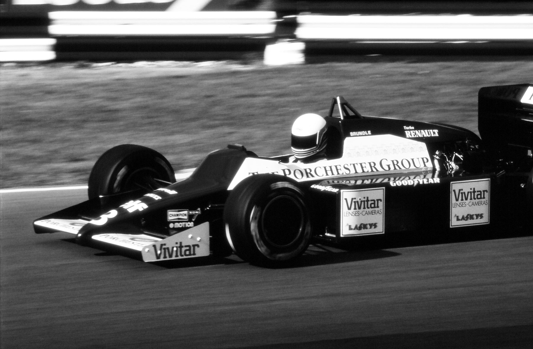 A dark single-seater with pale flanks lettered ‘Vivitar’ and ‘Porchester Group’ runs past the camera on a sunny circuit, the driver’s white helmet just above the cockpit.
