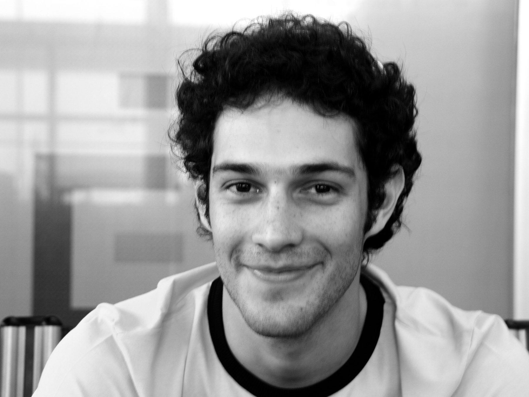 Head and shoulders of a smiling young man with thick, dark curly hair, in a pale T-shirt with a dark collar, photographed in a bright interior.