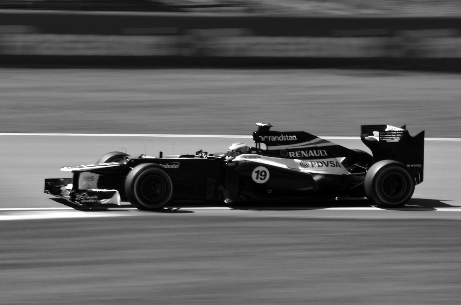 A dark Williams Formula One car, number 19, ‘Renault’ on its engine cover, is caught sharp against a streaked blur of grass and barrier in a panning shot.