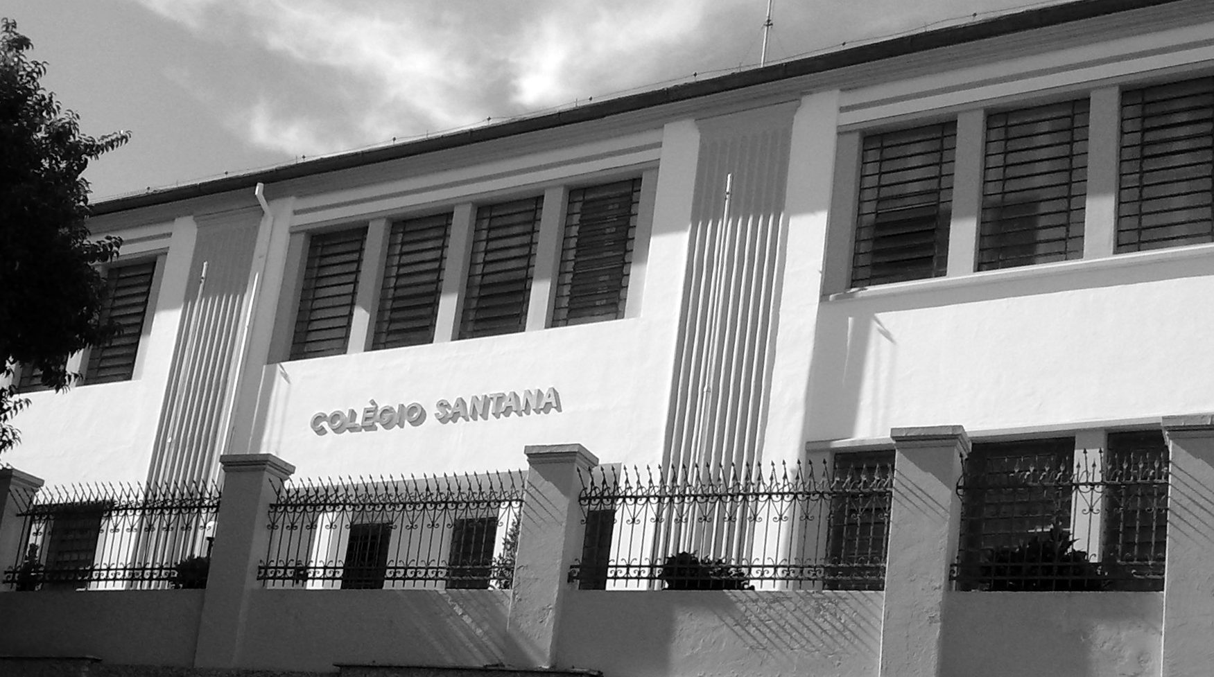 The long, pale façade of a school building with shuttered windows, the name Colégio Santana in raised letters on its wall, behind a low wall and iron railings.