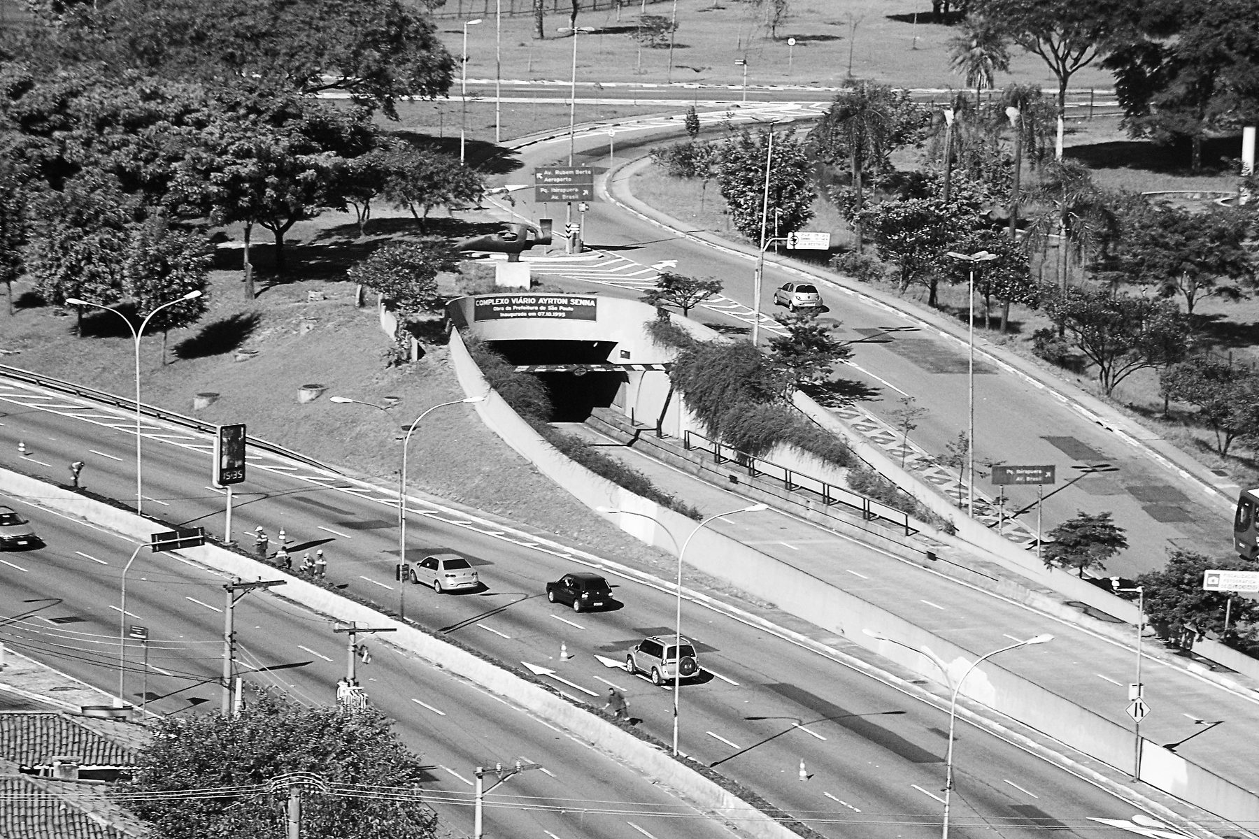 High view of a city avenue running into a tunnel whose portal bears the name ‘Complexo Viário Ayrton Senna’, with a bronze sculpture on a plinth above it.