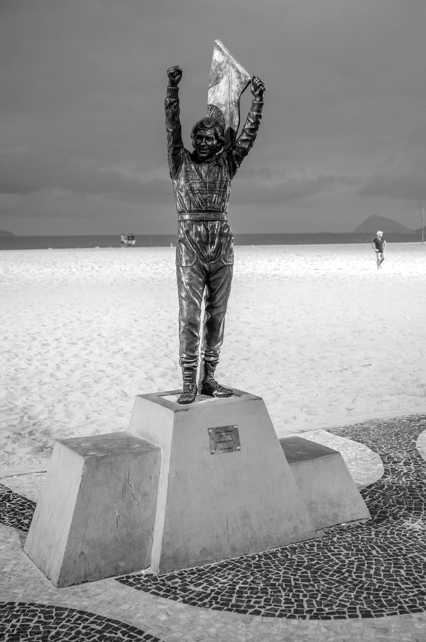 Al atardecer, la estatua de bronce de un piloto con los dos brazos en alto, sosteniendo una bandera, se alza sobre un pedestal de tres escalones en el pavimento con dibujo de ondas de un paseo marítimo, con el mar detrás.