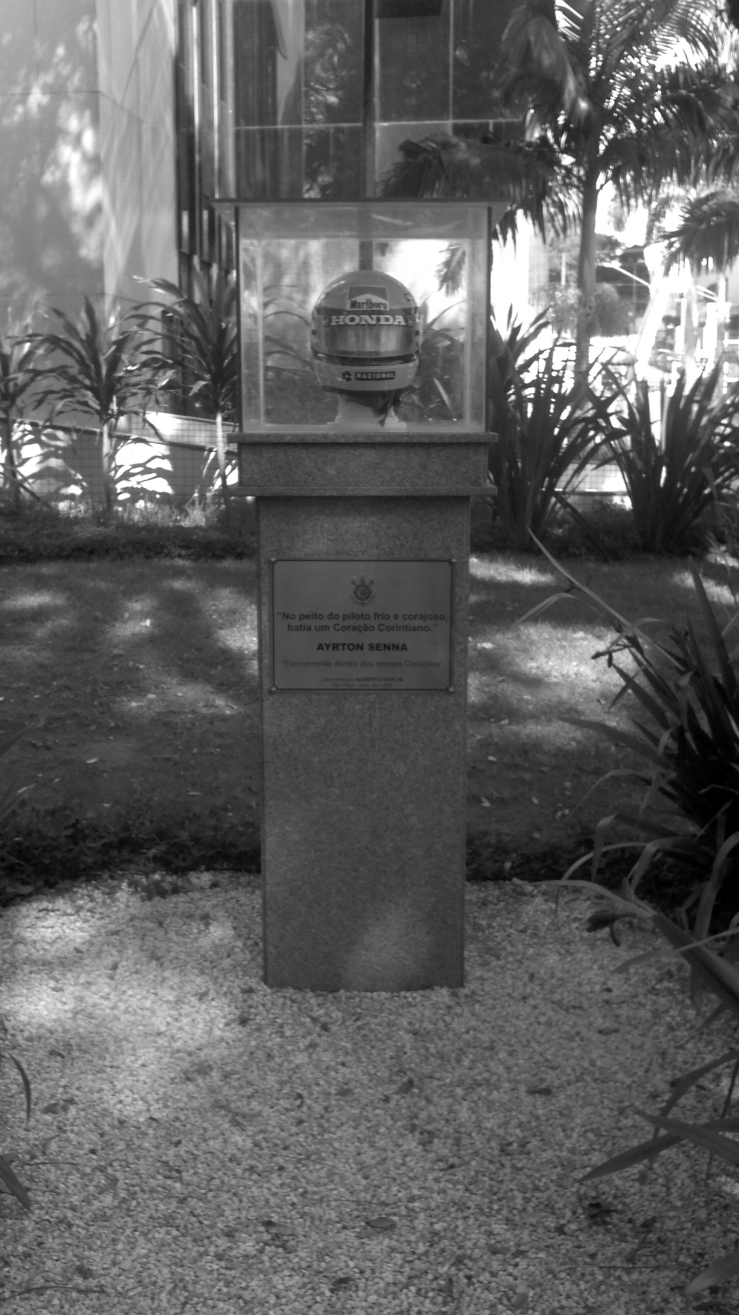 Un casco sobre un soporte blanco, dentro de una vitrina, corona un pedestal de granito entre palmeras, junto a la fachada acristalada de un edificio del club; una placa de acero en el pedestal lleva el escudo del club y un homenaje a Ayrton Senna.