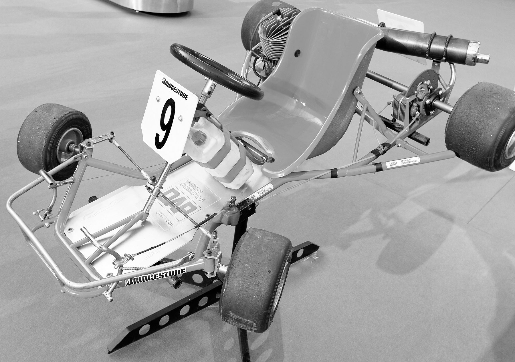 A racing kart on a display stand, seen from the front three-quarters: a tubular frame, a moulded seat, a steering wheel, a pale number board with a black 9, and an exposed engine behind the seat.