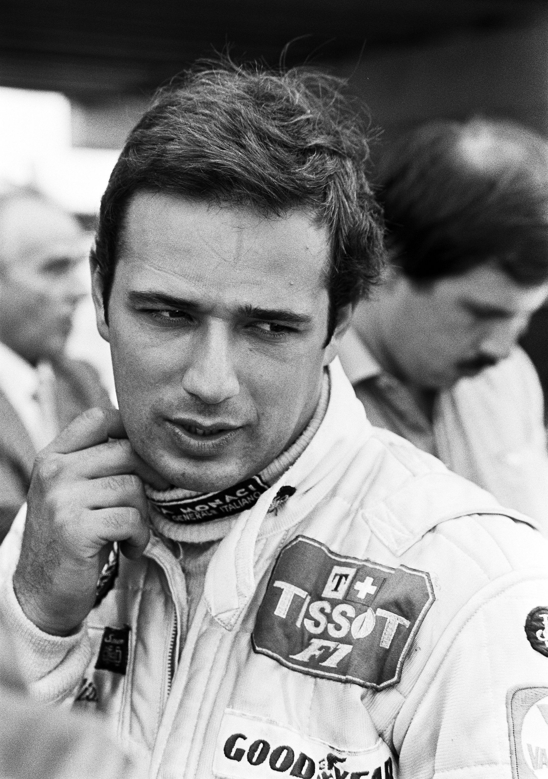 Portrait of a dark-haired young driver in white racing overalls, his hand raised to his chin, looking down to one side, with people behind him in the paddock.