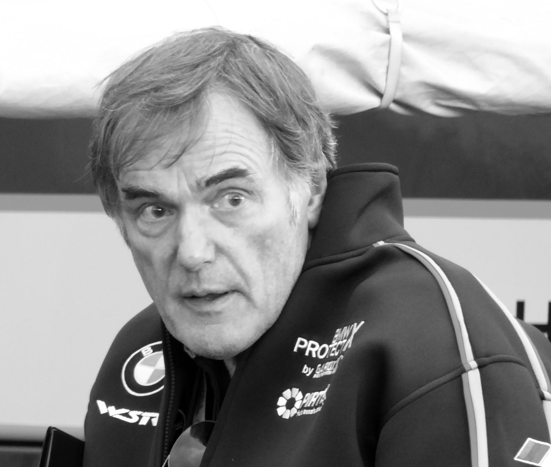A grey-haired man in a dark team jacket with a pale stripe down the sleeve looks up, mouth slightly open, in the pit lane.