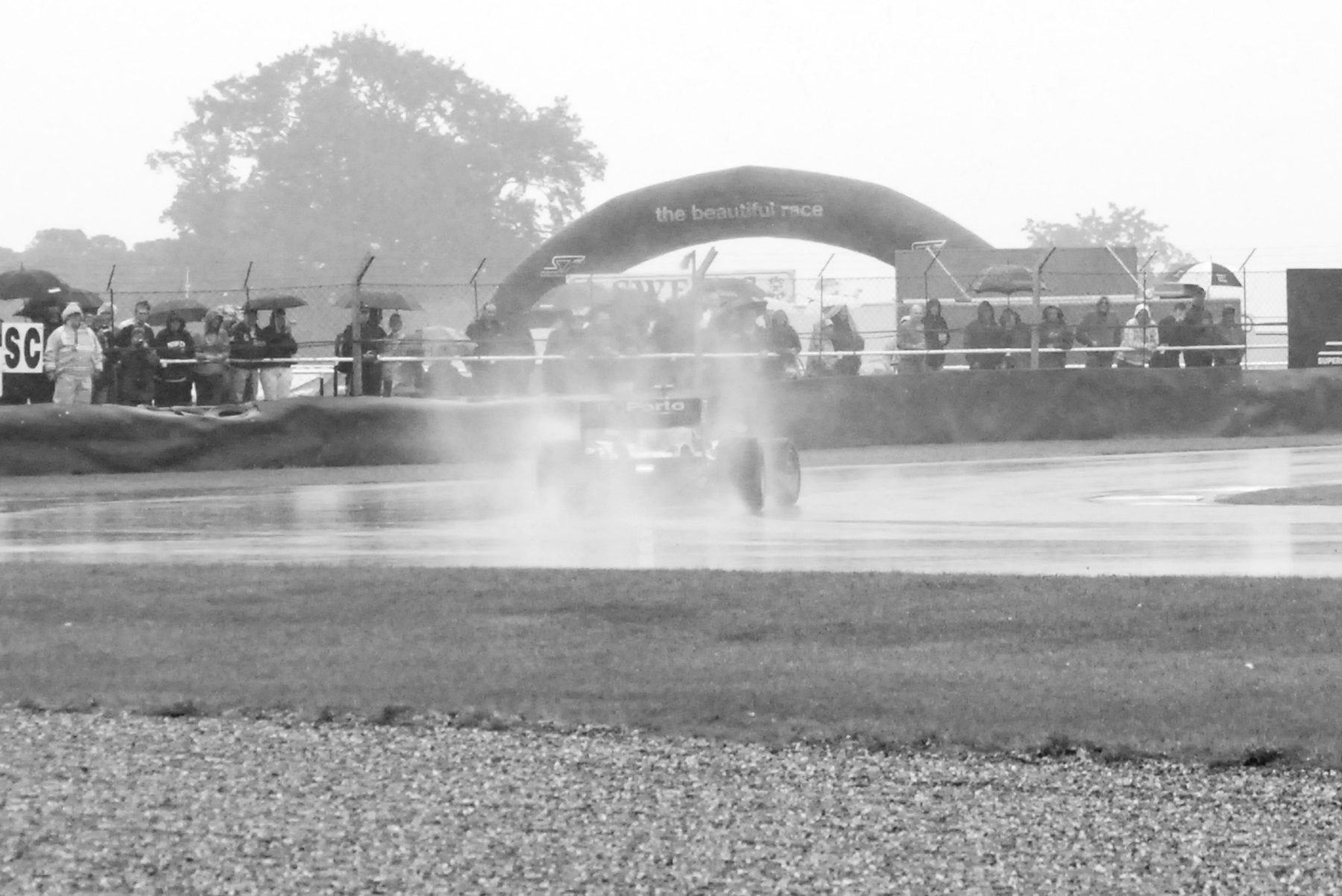 A single-seater throws up a plume of spray on a rain-soaked circuit, blurred by rain and mist, with a banner arch, spectators and trees faint behind.