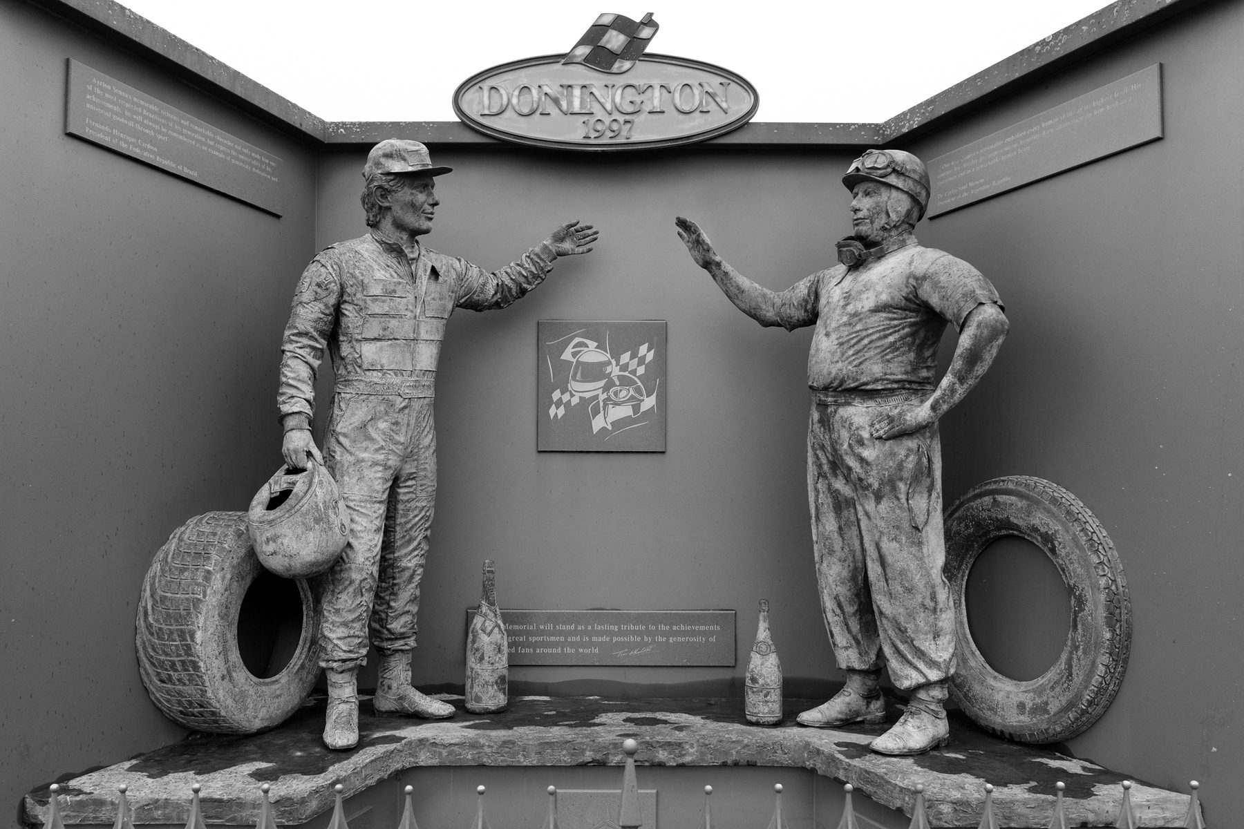 A memorial of two bronze figures in racing overalls facing each other in front of a dark wall, one holding a helmet, the other gesturing, each with a tyre beside him and a champagne bottle at his feet, under a sign reading ‘Donington’.