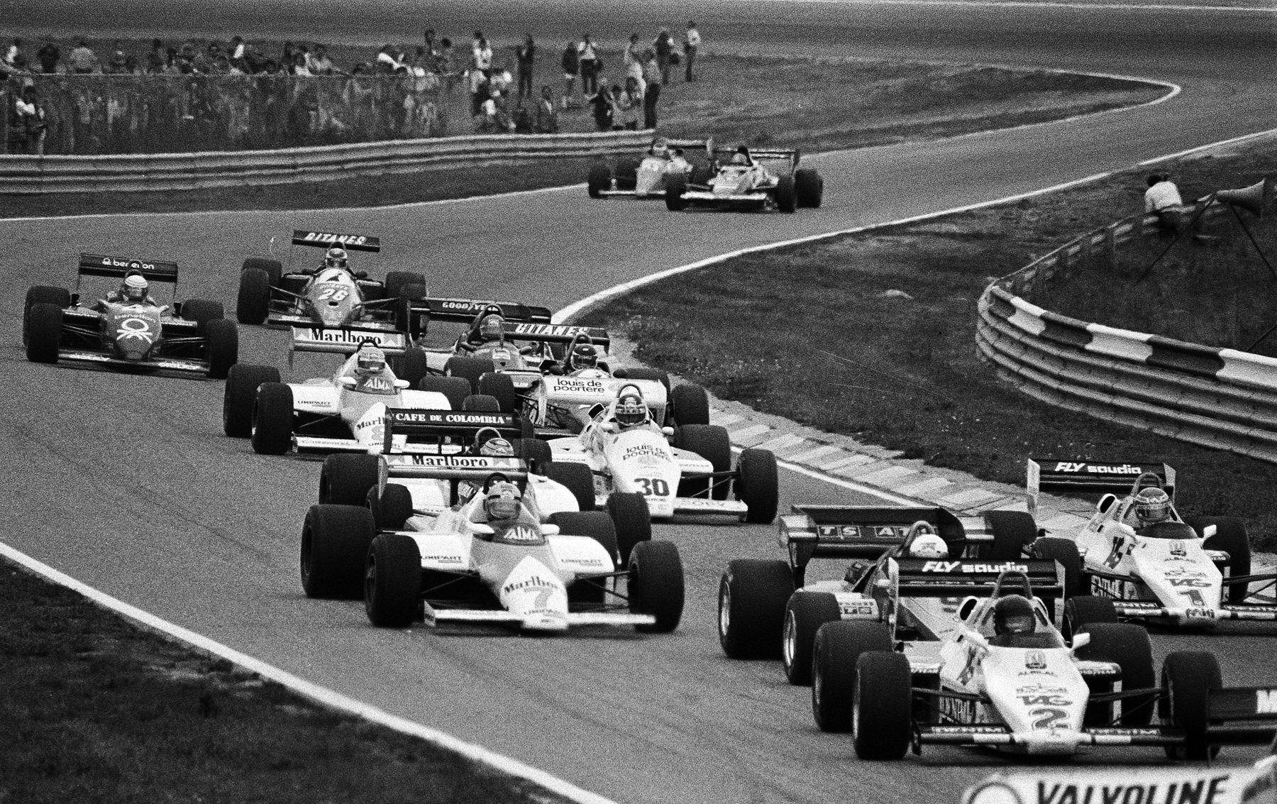 A pack of Formula One cars streams through a long curve, the car numbered 2 nearest the camera at right, number 1 behind it, spectators lining the bank on the far side.