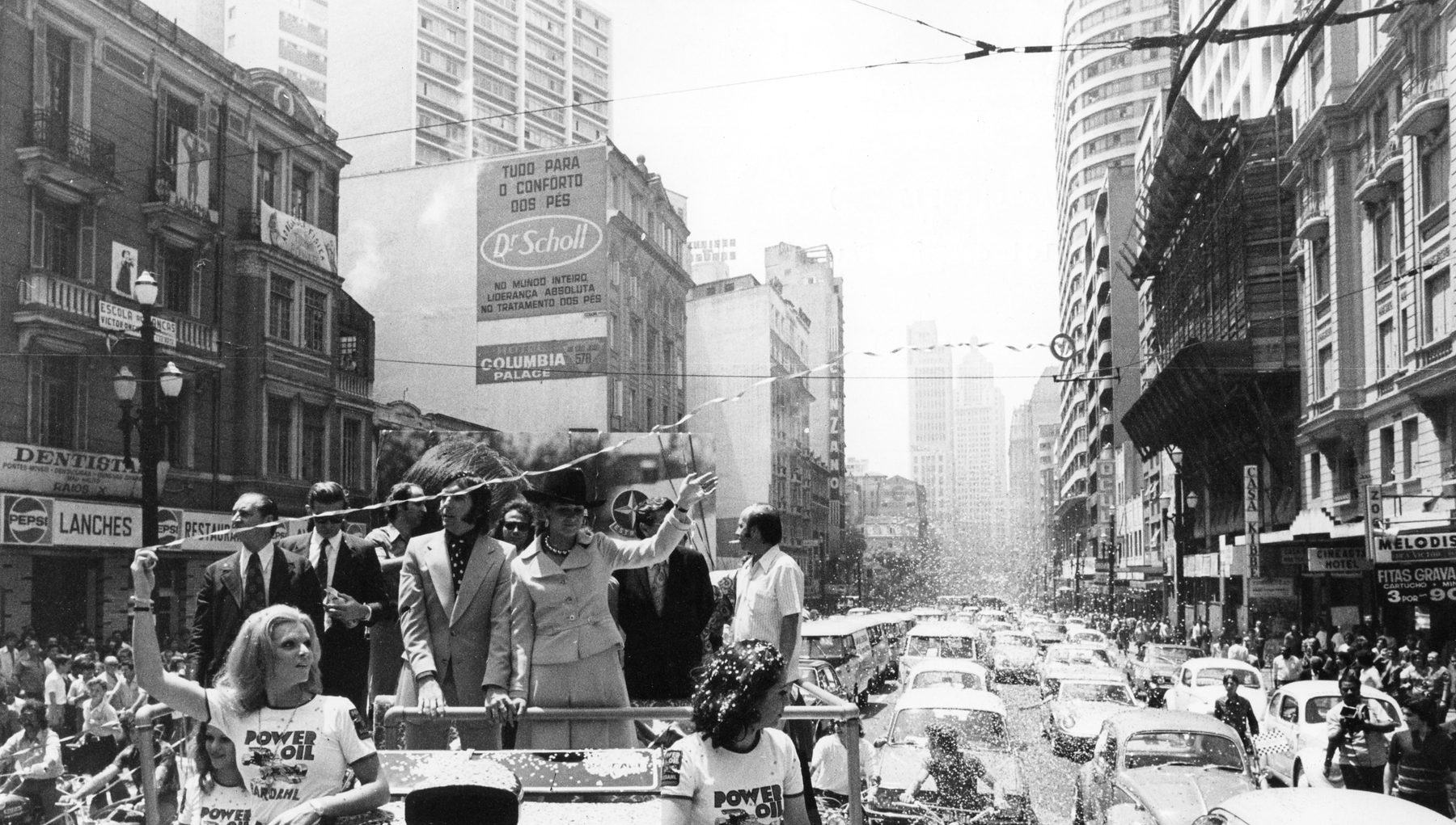 Men in suits and a woman in a pale suit and dark hat stand waving on an open vehicle as confetti falls on a crowded city avenue lined with tall buildings, cars and motorcycles; young women in sponsors’ T-shirts ride at the front, one waving.