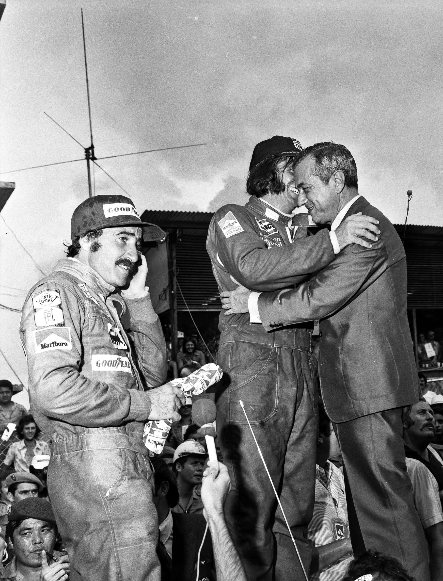 On a crowded podium a man in a suit embraces a driver in racing overalls and cap; another moustached driver in overalls and cap stands at left with a bouquet, a hand to his ear.