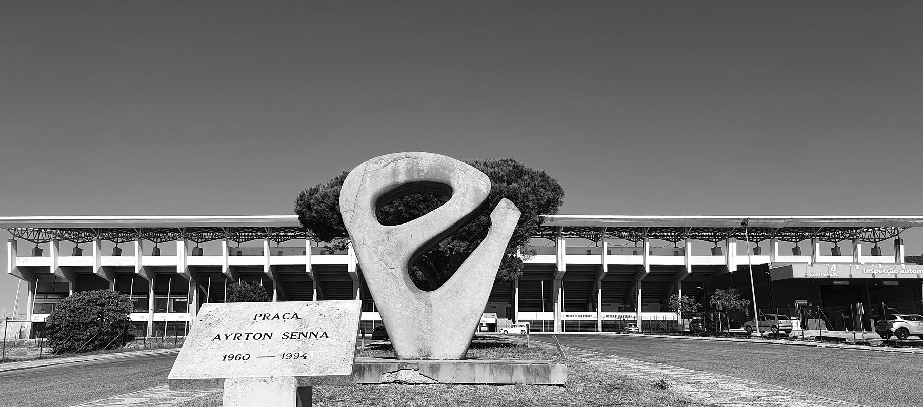 A roundabout garden with a small stone marker reading ‘Praça Ayrton Senna, 1960–1994’ and a tall abstract stone sculpture shaped like a looping S, the circuit’s long white grandstand behind.