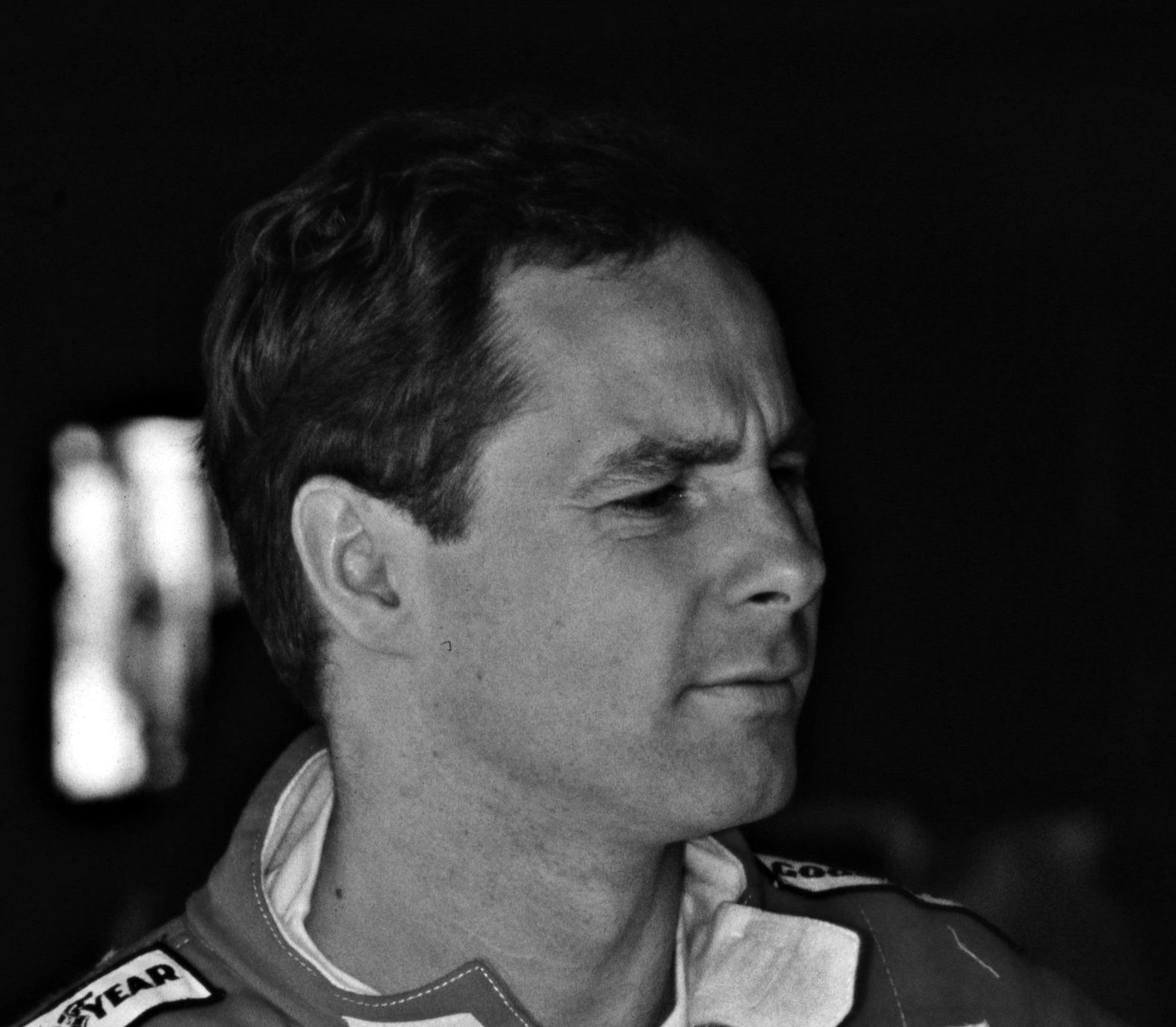 Head and shoulders of a dark-haired driver in team overalls, collar open, listening intently to someone out of frame against a dark background.