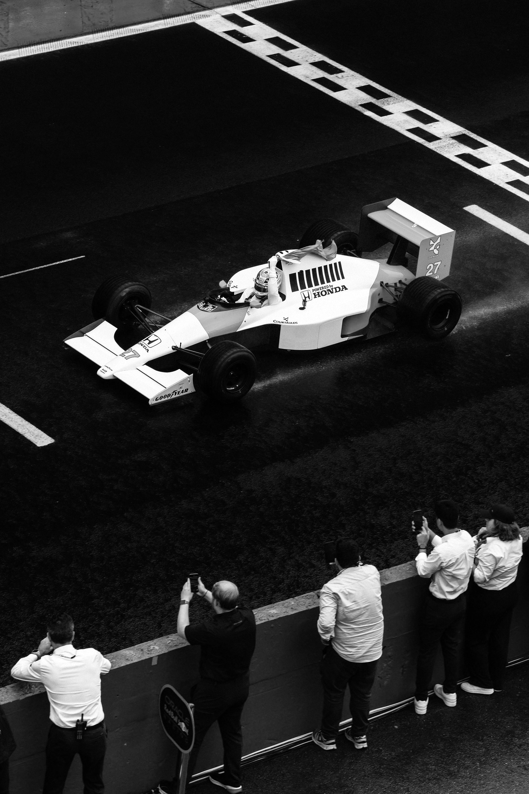 Looking down on a wet start line: a pale McLaren numbered 27 crosses the chequered stripe, a flag held up from the cockpit, while team staff and guests follow it with phones from the pit wall below.