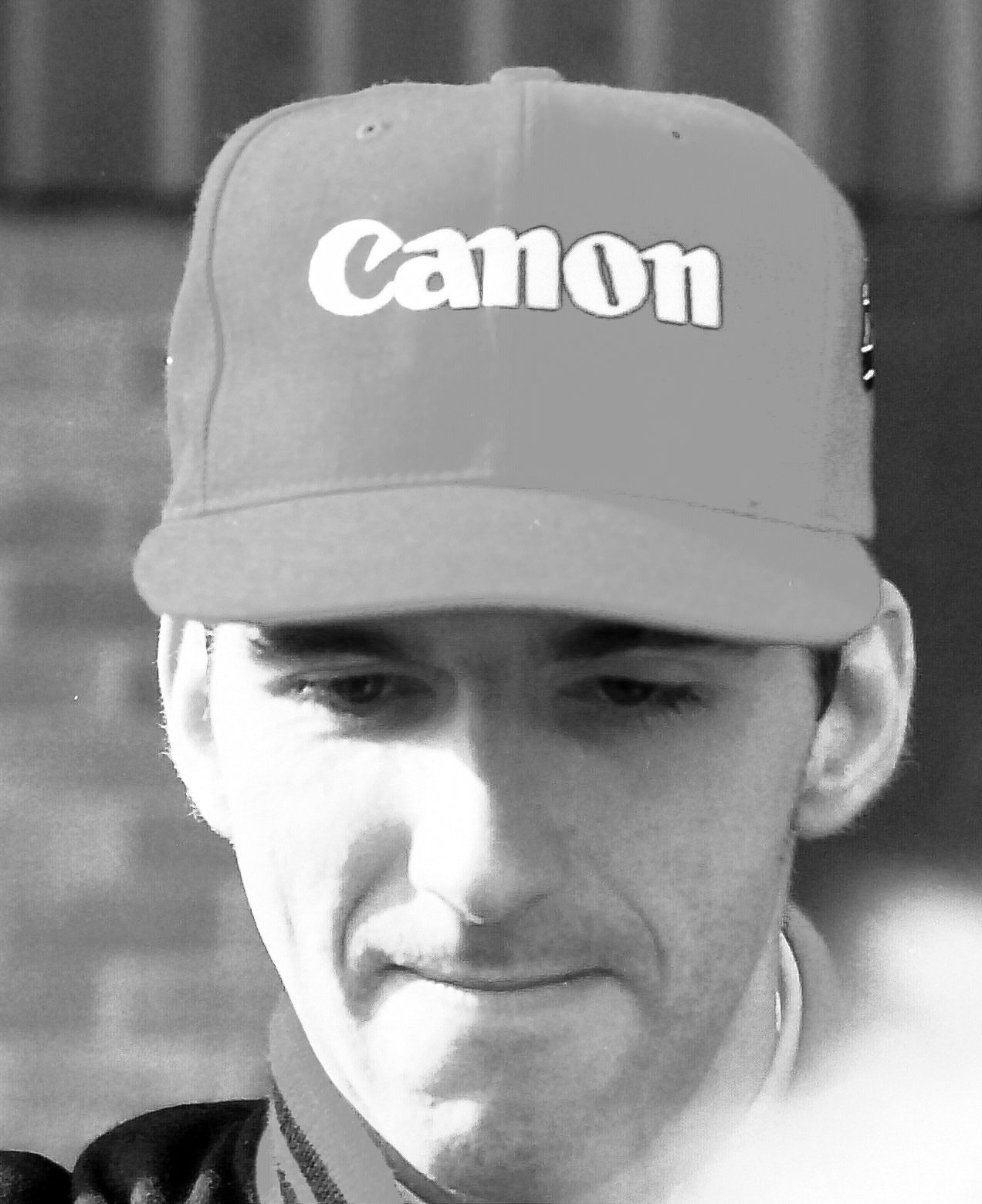 A dark-haired driver in a baseball cap lettered ‘Canon’ looks down, lips pressed together, his eyes shaded by the peak.