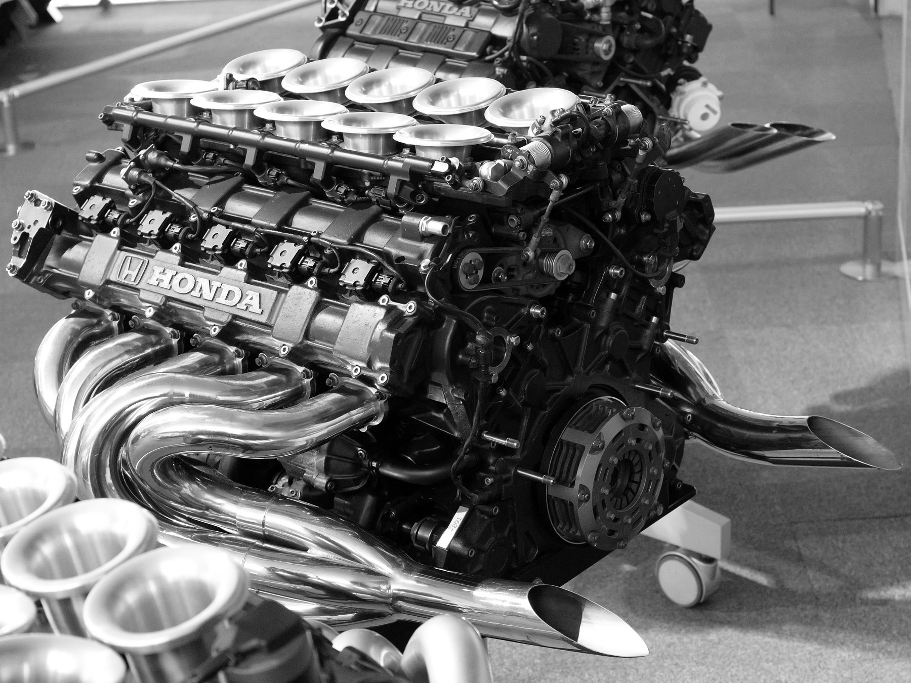 A racing engine on a display stand: a row of polished intake trumpets above black cam covers marked HONDA, and chrome exhaust pipes curling down to two tailpipes; the out-of-focus trumpets of a second engine fill the lower left corner.