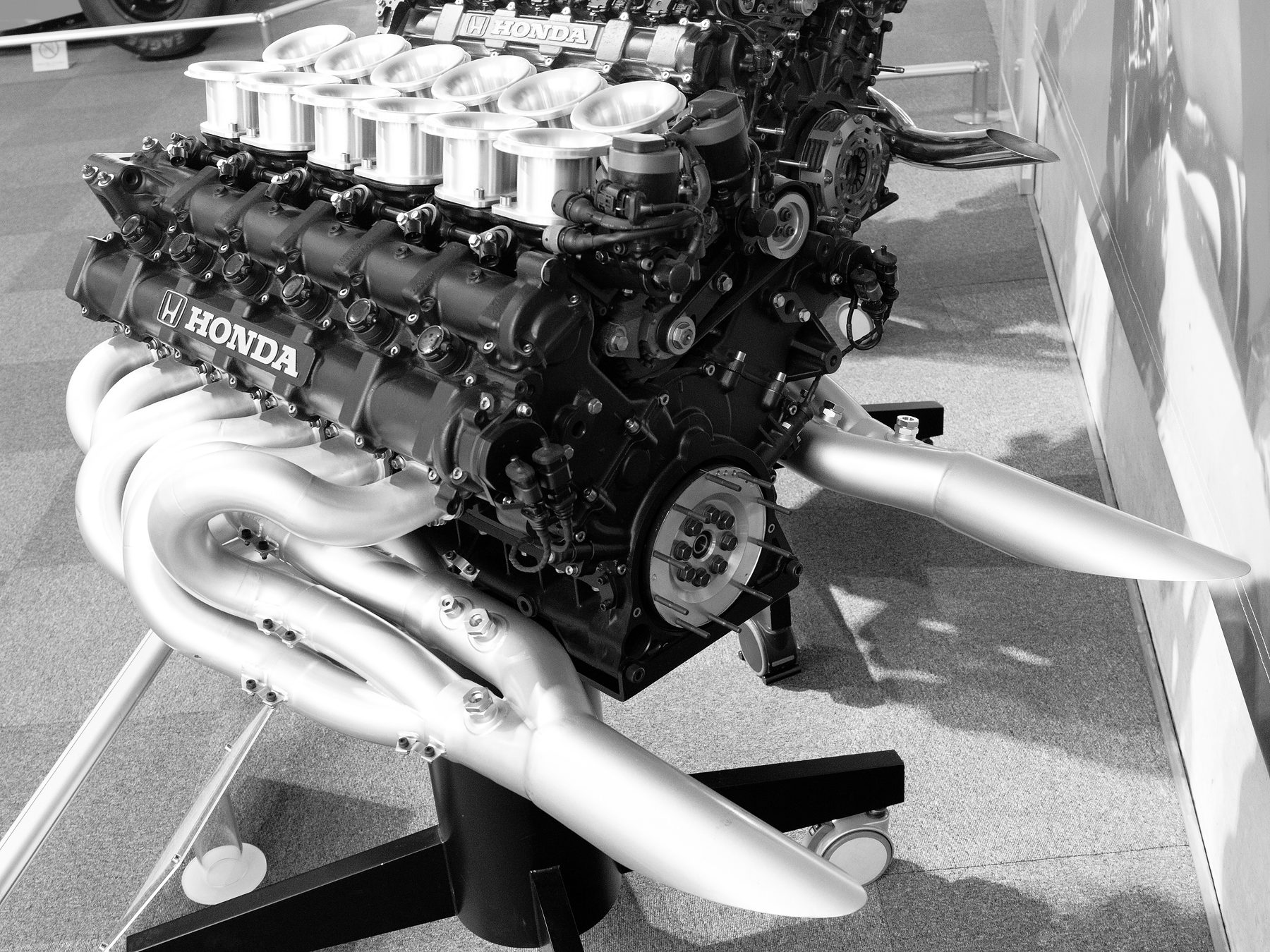 A racing engine on a black display stand: 12 polished intake trumpets above black heads marked HONDA, and pale exhaust pipes sweeping back into two long tailpipes.