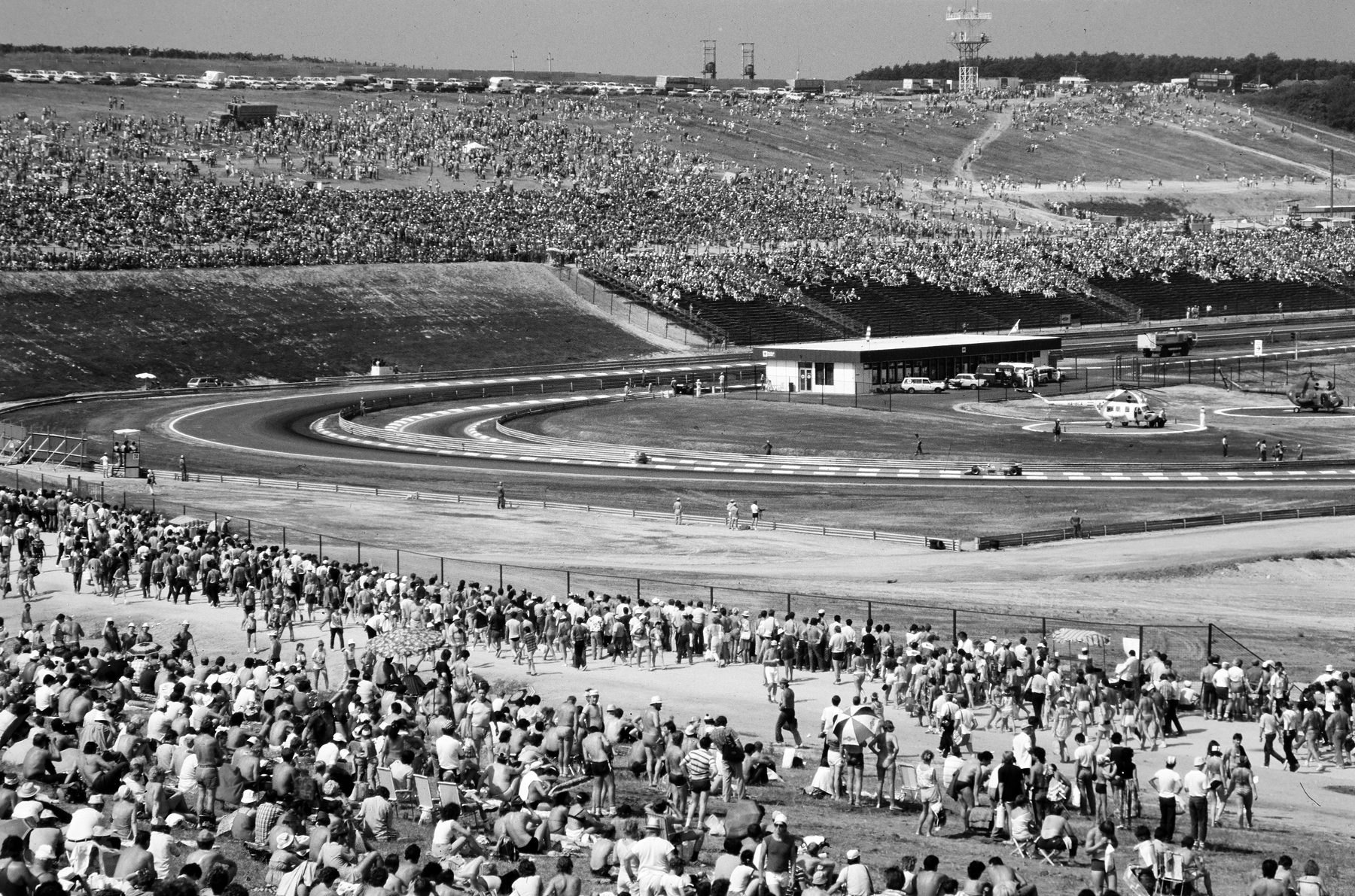 A huge crowd covers the dry grass banks overlooking a new circuit in a shallow valley, with more spectators on the far hillside and the pit buildings in the distance.