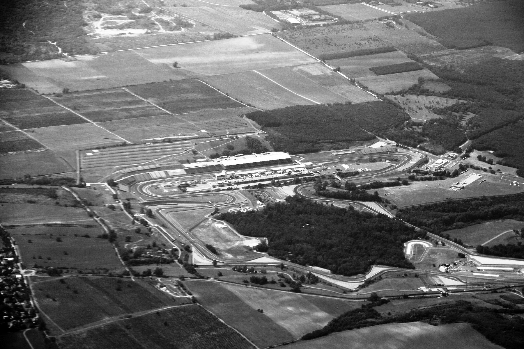 An aerial view of a twisting circuit set among fields and woodland, its pit buildings and grandstands along one straight, softened by haze.