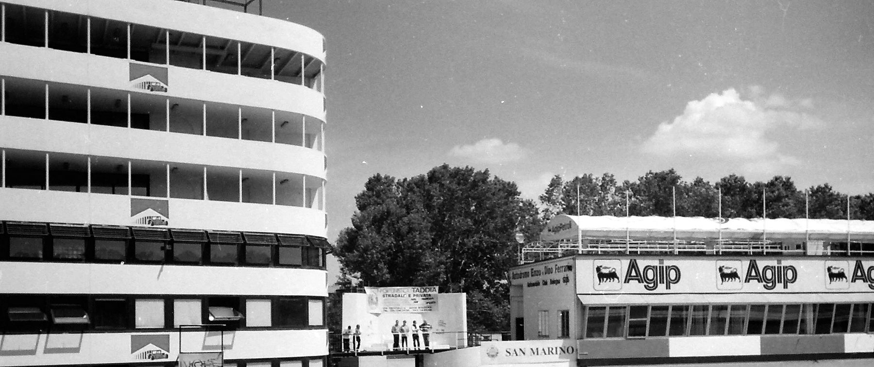 A tall, curved white control tower beside a small podium and the long pit building, its roof banner lettered with a fuel company’s name, under a summer sky.