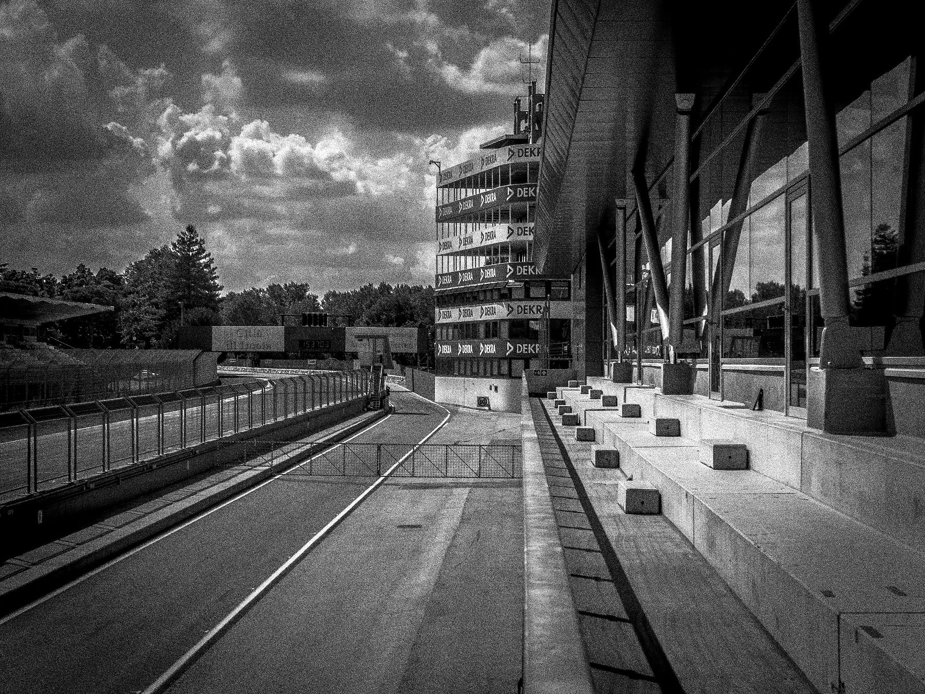 An empty pit lane runs between the garages and the pit wall towards a tall control tower under dark, heaped storm clouds.