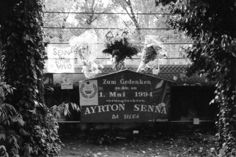 A dark memorial board with lettering and a small wreath emblem stands against ivy-covered railings, bunches of flowers tied beside it and a hand-painted sign above.
