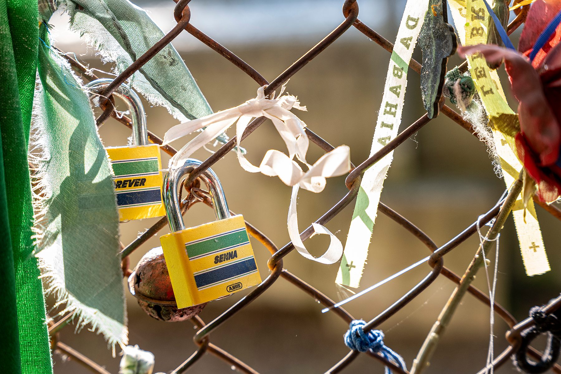 Close-up of padlocks painted yellow with green and blue bands, like his helmet, locked to a chain-link fence among faded ribbons and scraps of cloth.