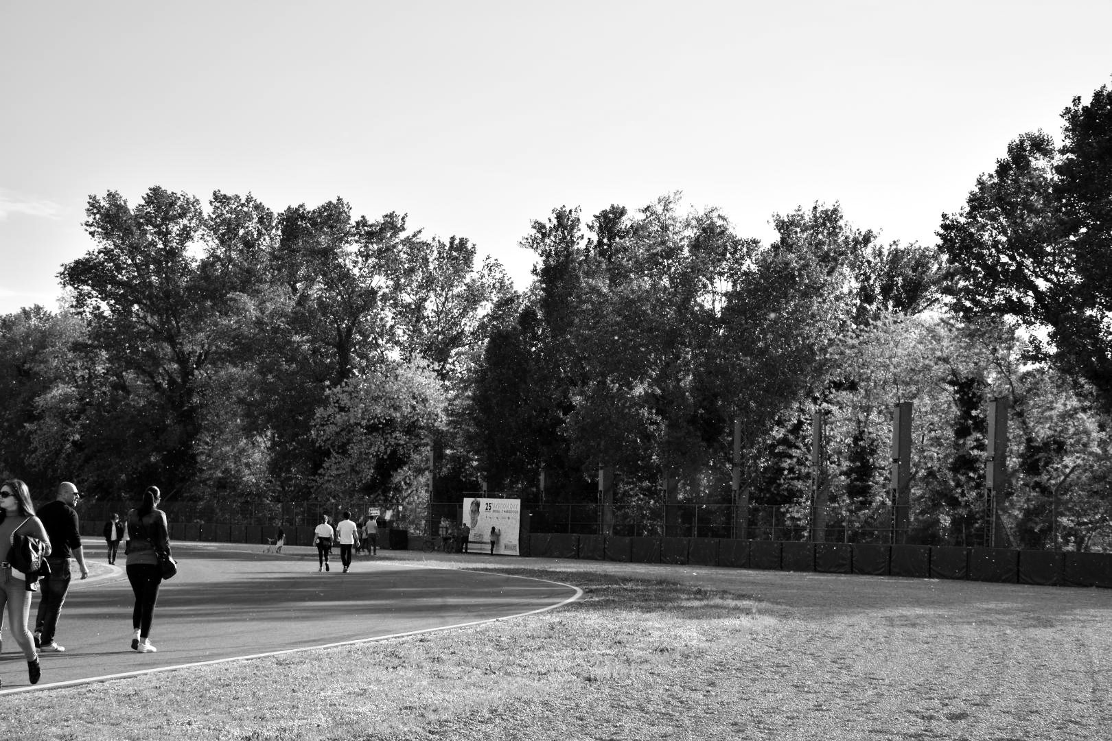 People stroll along an empty circuit as it curves past a grass verge, a fence and a line of tall trees; by the fence stands a board bearing his portrait.