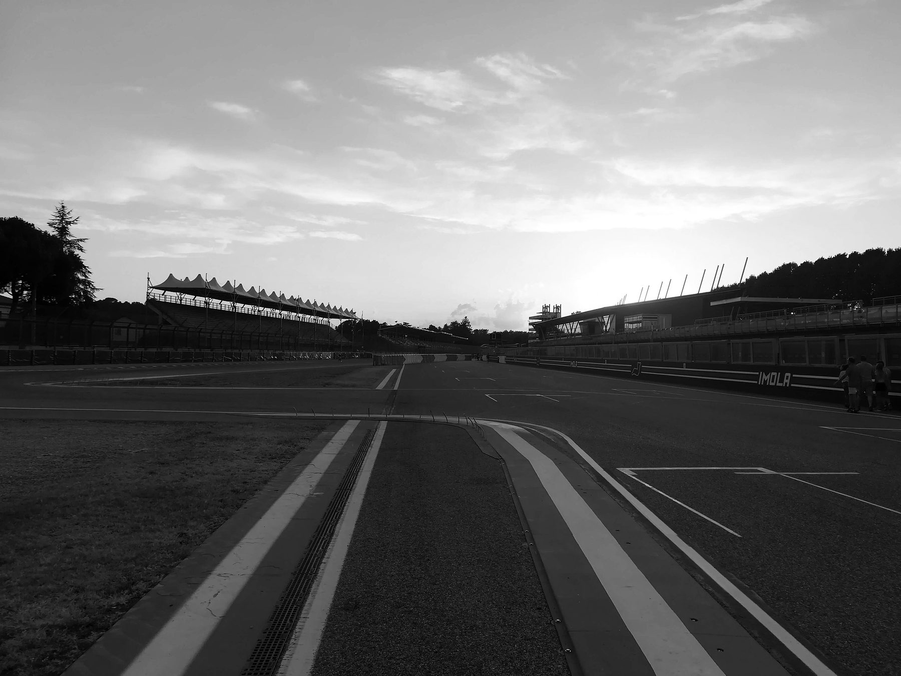 Striped kerbs lead the eye down an empty straight towards the grandstands and the low sun under a streaked evening sky; four people stand far off by the pit wall at the right.