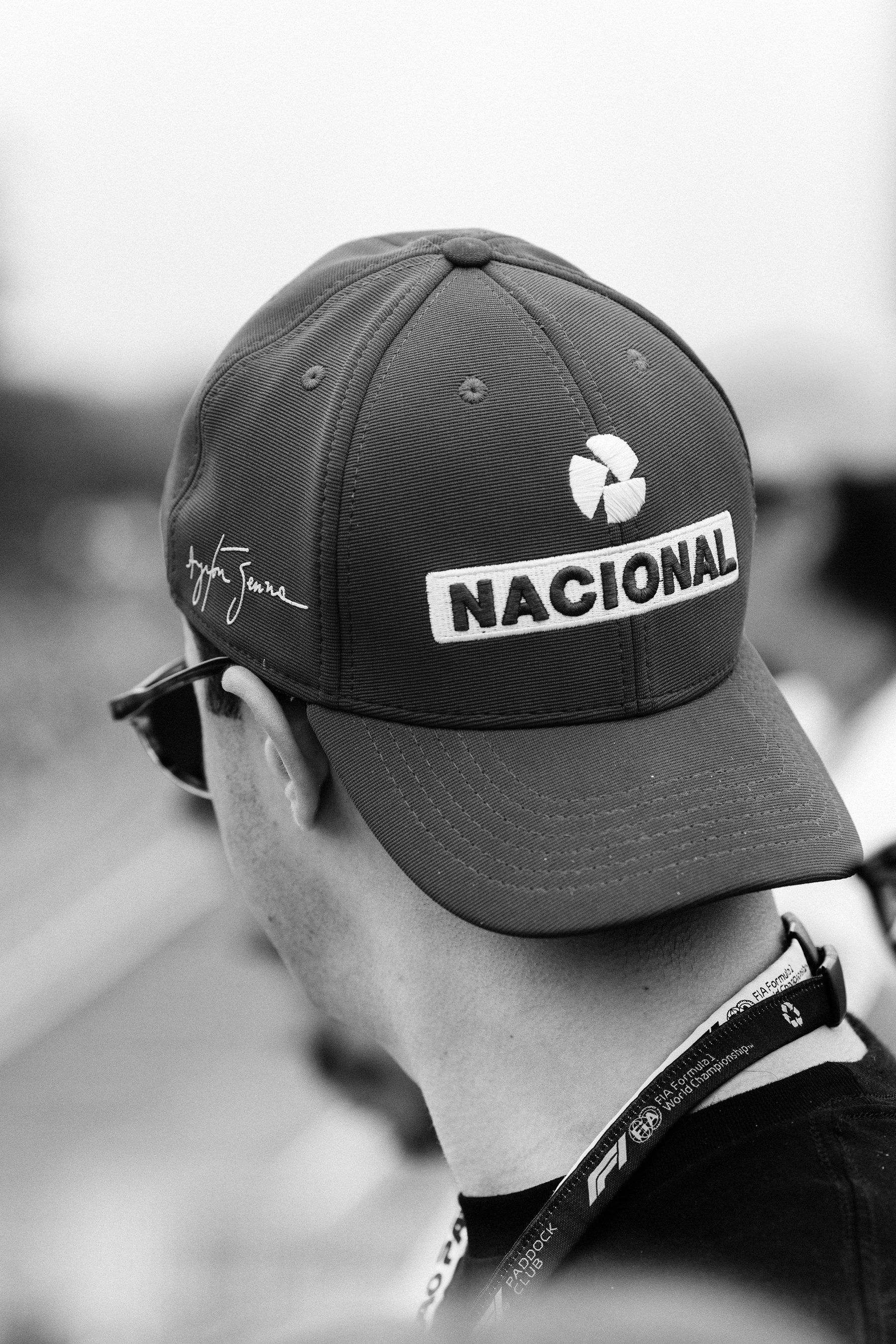 Seen from behind and to one side, a man in sunglasses wears a dark cap with a pale ‘Nacional’ badge and a looping signature embroidered on the side.