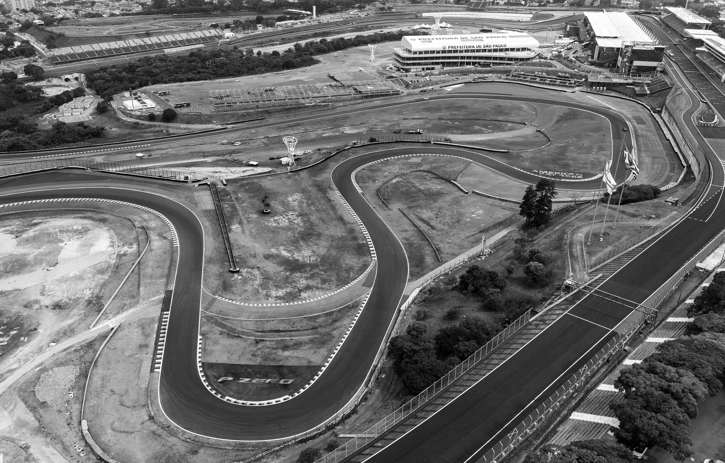 Vista aérea de la sinuosa zona interior de un circuito, con curvas y rectas cortas entre la hierba, los boxes y las tribunas arriba y la ciudad al fondo.