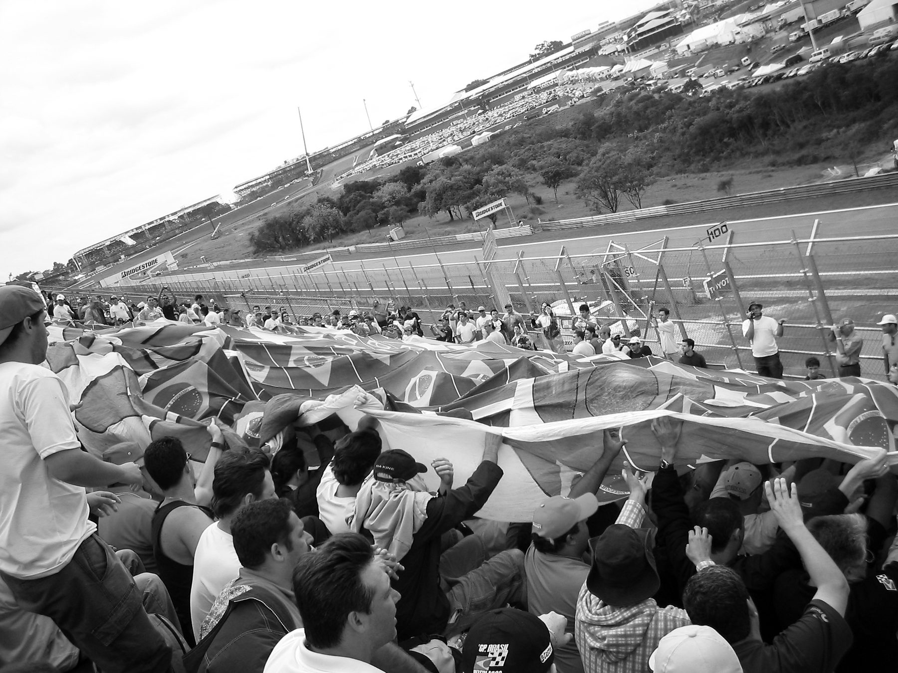 A packed grandstand crowd reaches up to pass a huge patchwork flag over their heads, the track and a hillside beyond.