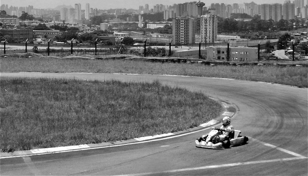 A lone kart rounds a bend edged with painted kerbs on a narrow track set in rough grass, with the tower blocks of a city on the horizon.