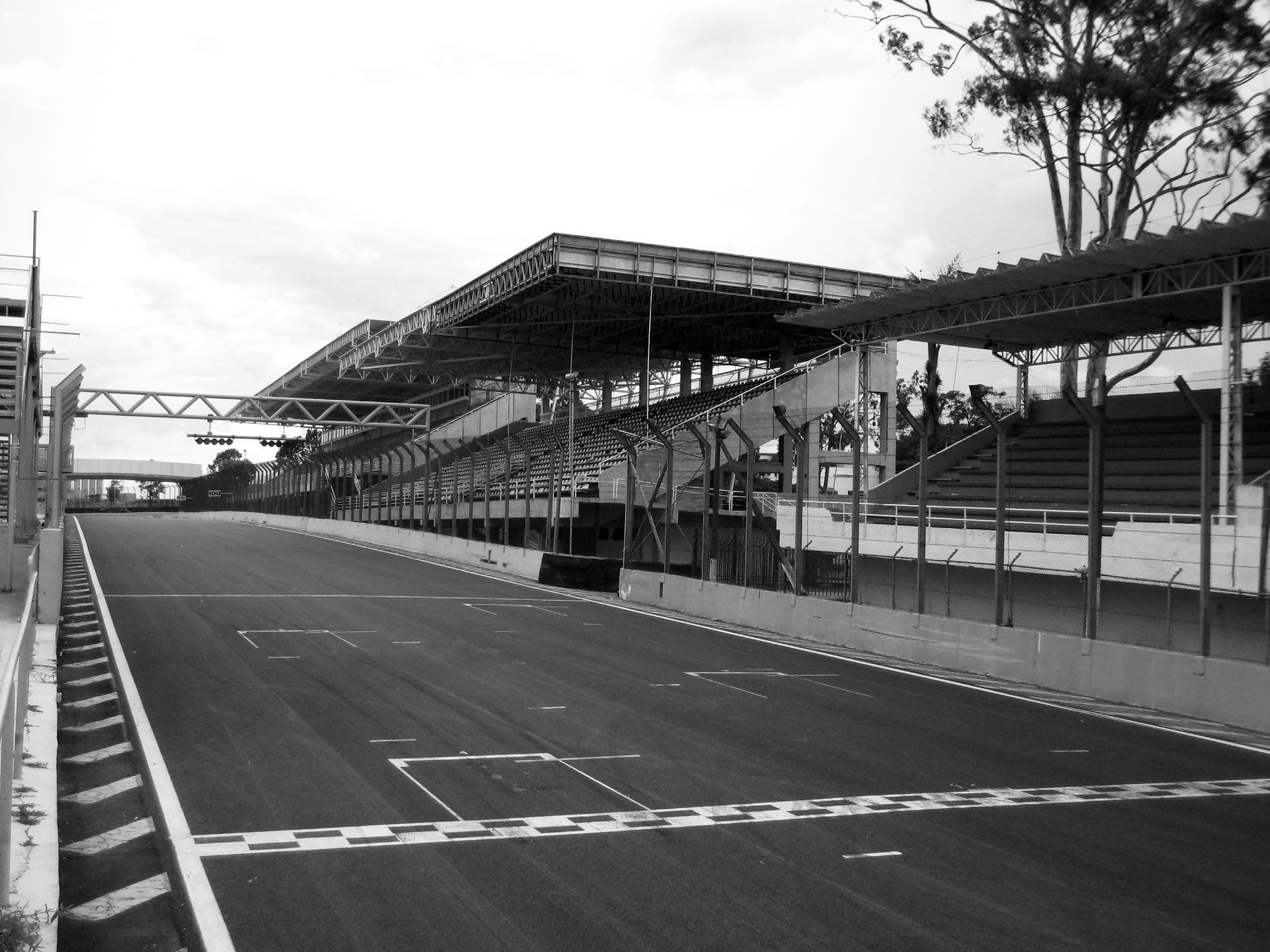 An empty start-finish straight with painted grid boxes runs past a long, covered grandstand and pit buildings under a pale sky.