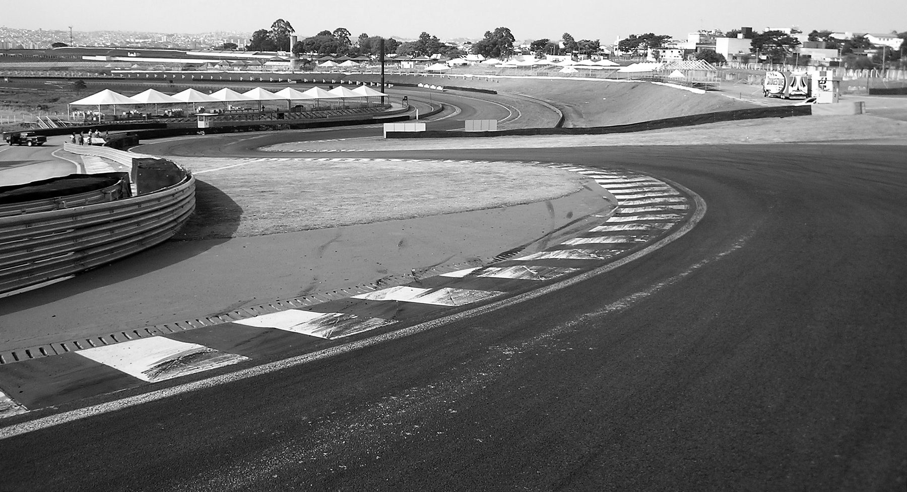 From the edge of the track, a striped kerb curves round a run-off area as the circuit winds downhill and away through a sequence of bends towards distant white tents.