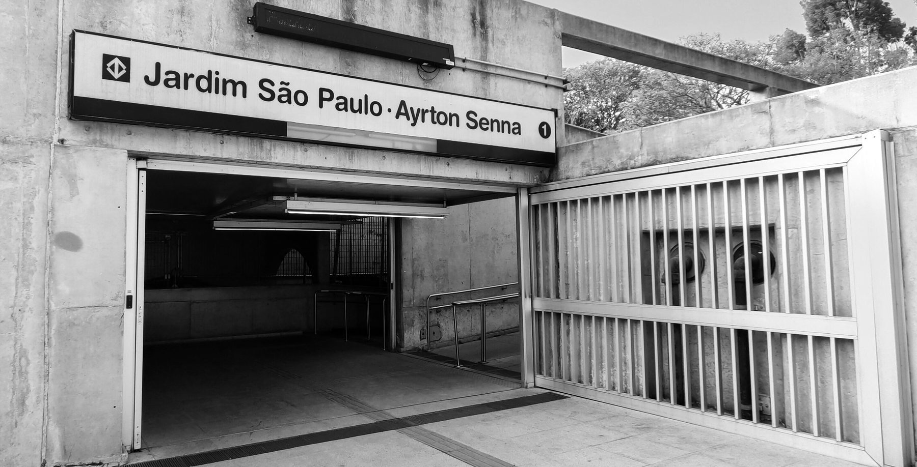 The concrete entrance to a metro station under a sign reading ‘Jardim São Paulo–Ayrton Senna’, its doorway open and its gate folded back.