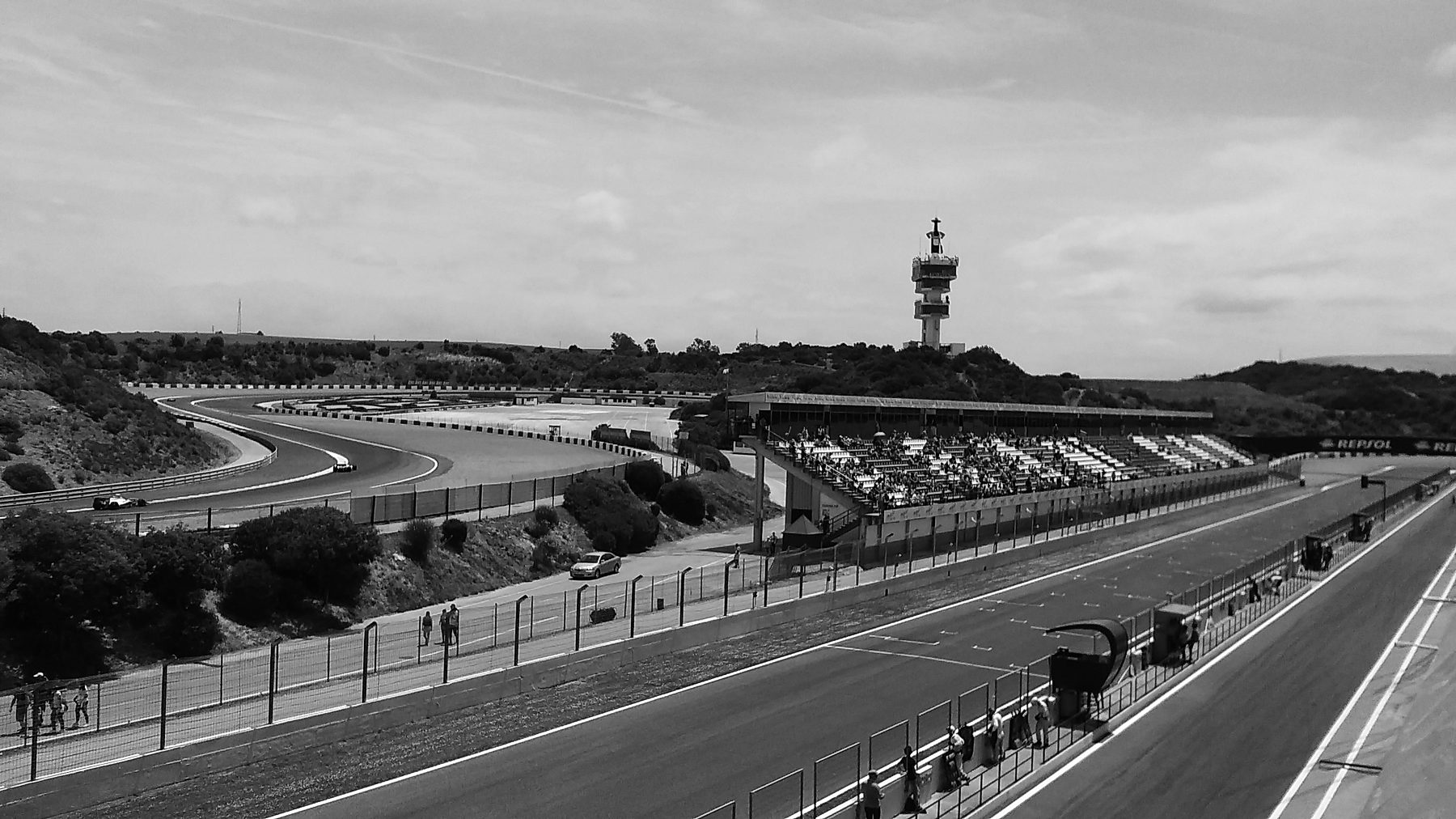 A long pit straight bordered by a grandstand runs towards a slender control tower on a dry hillside, with the circuit winding through scrub beyond.