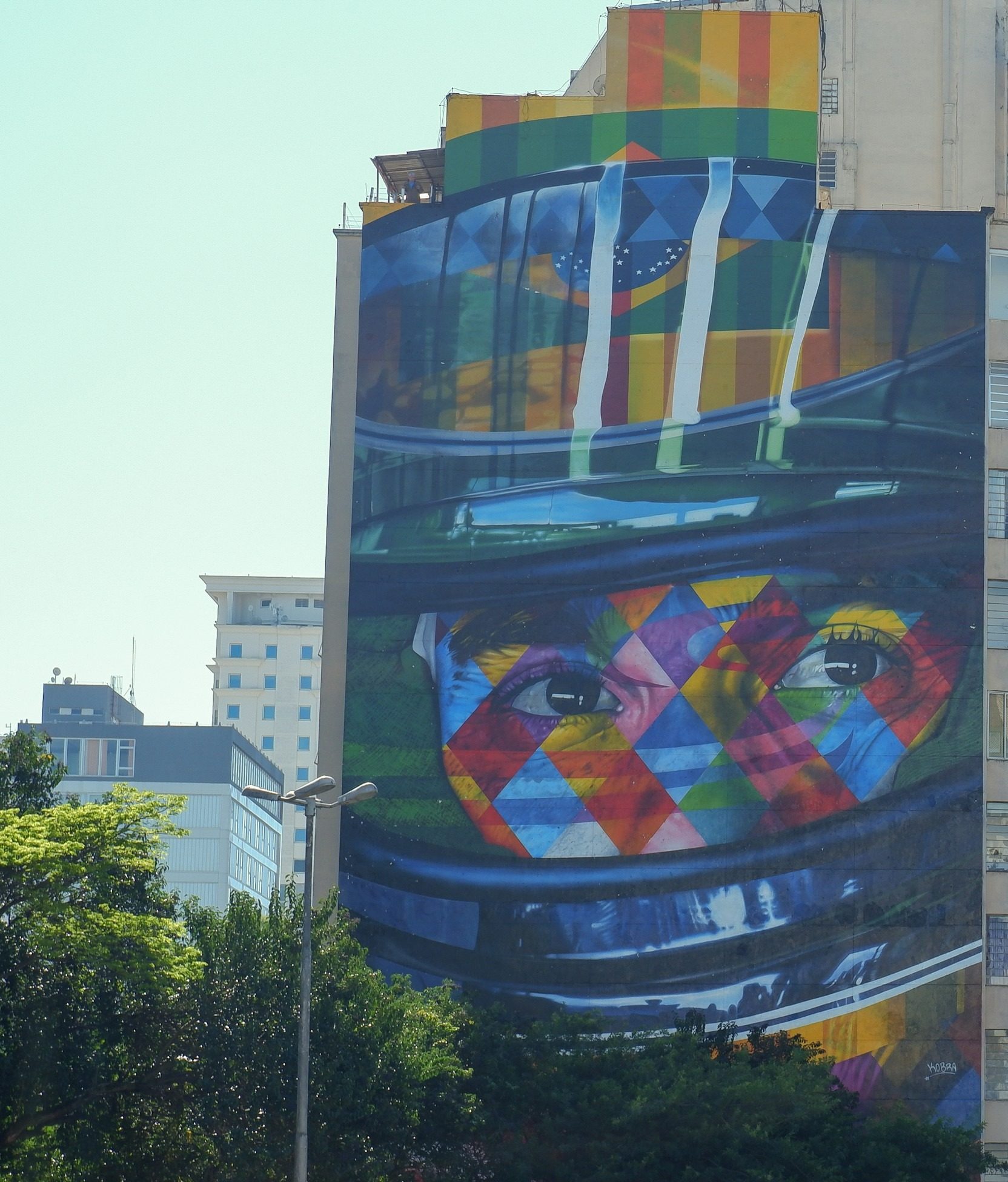 The whole gable of a tall city building is painted with a huge, brightly patterned portrait of a driver’s eyes looking out through the open visor of a helmet, a Brazilian flag reflected in it, above a line of trees and a street lamp.