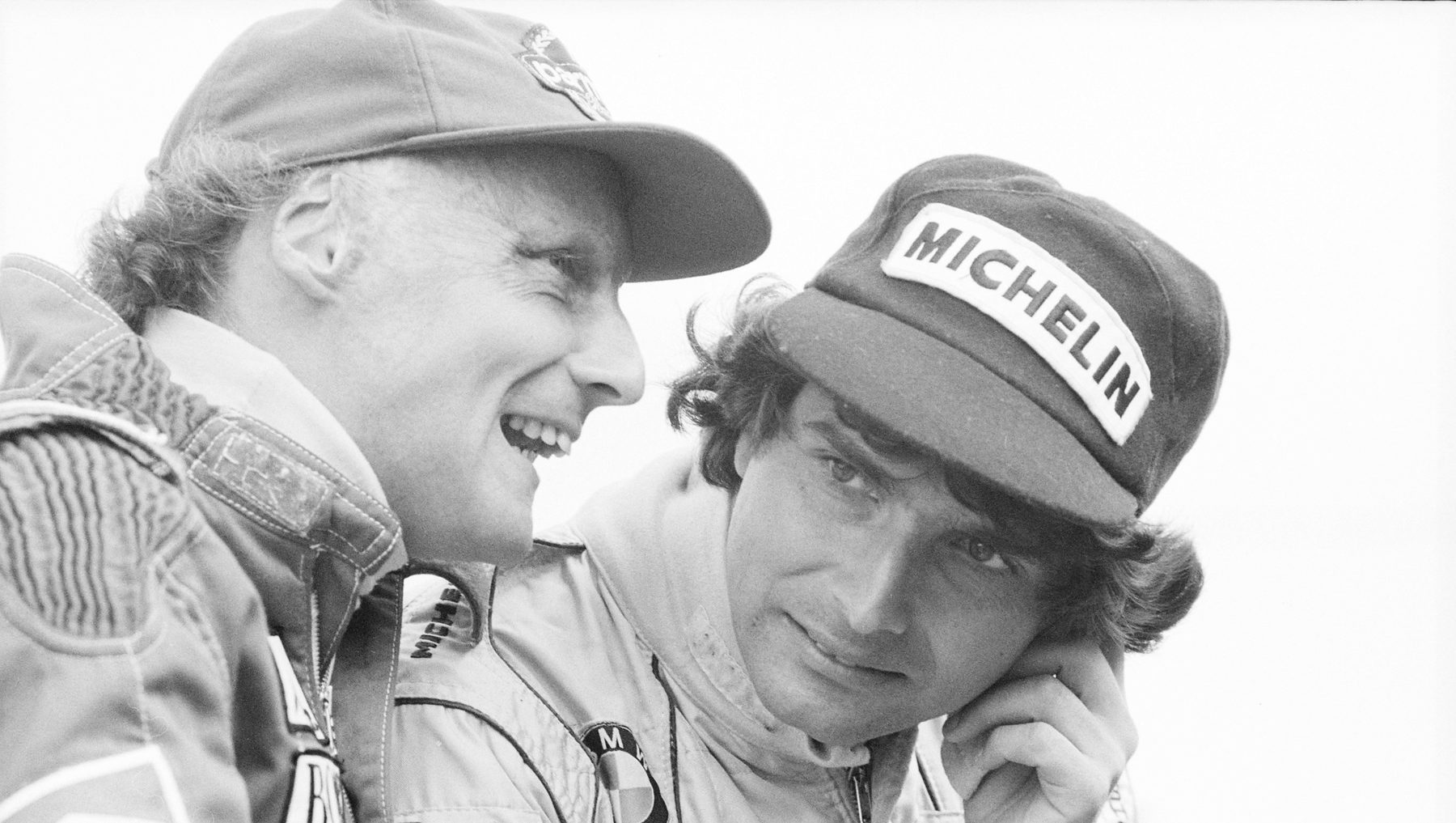Close-up of two drivers in caps and overalls sharing a joke, one grinning broadly, the other listening with a hand to his ear.