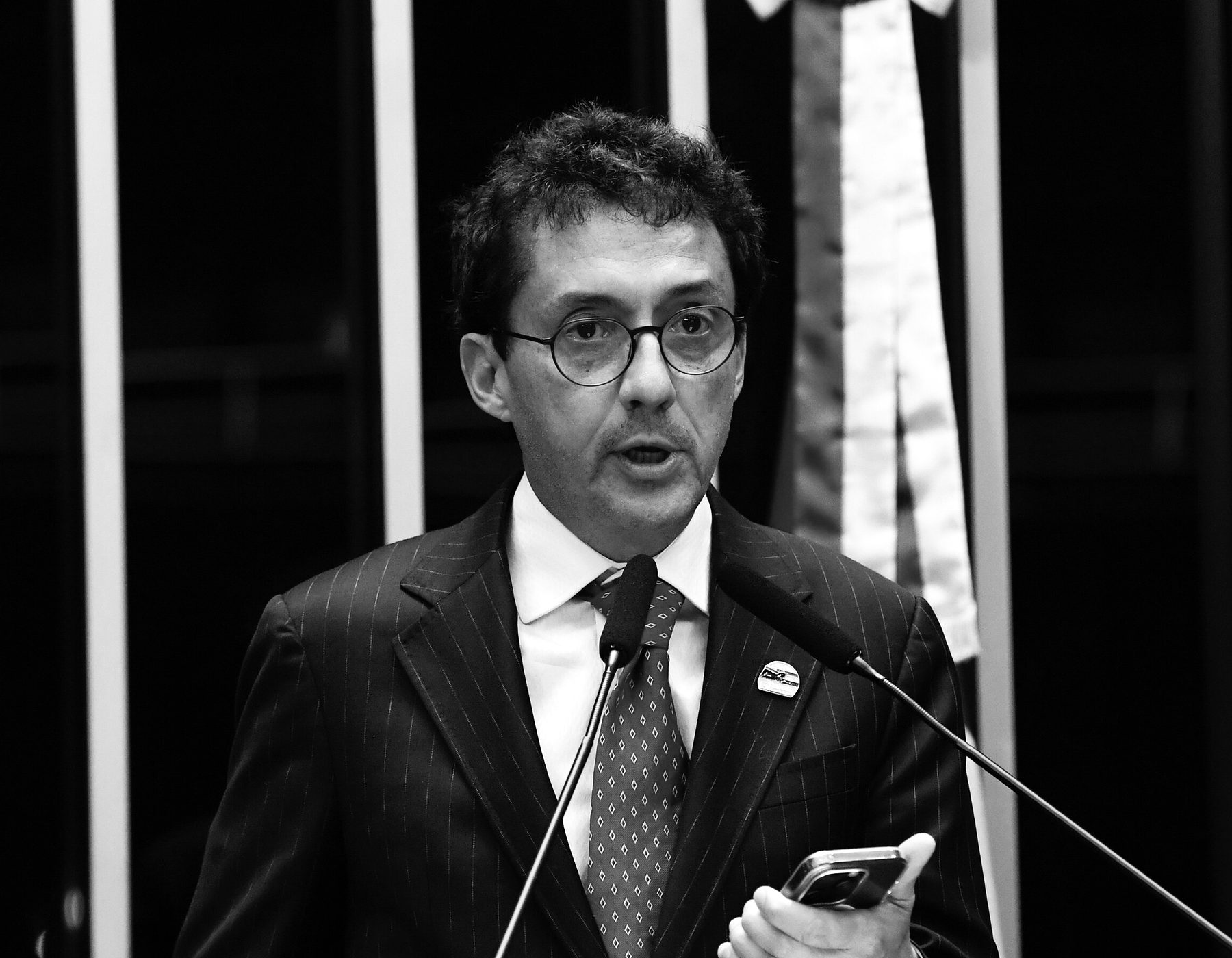 A man with dark, wavy hair and round glasses, in a pinstriped suit, speaks at a microphone holding a phone, a banded sash hanging behind him.